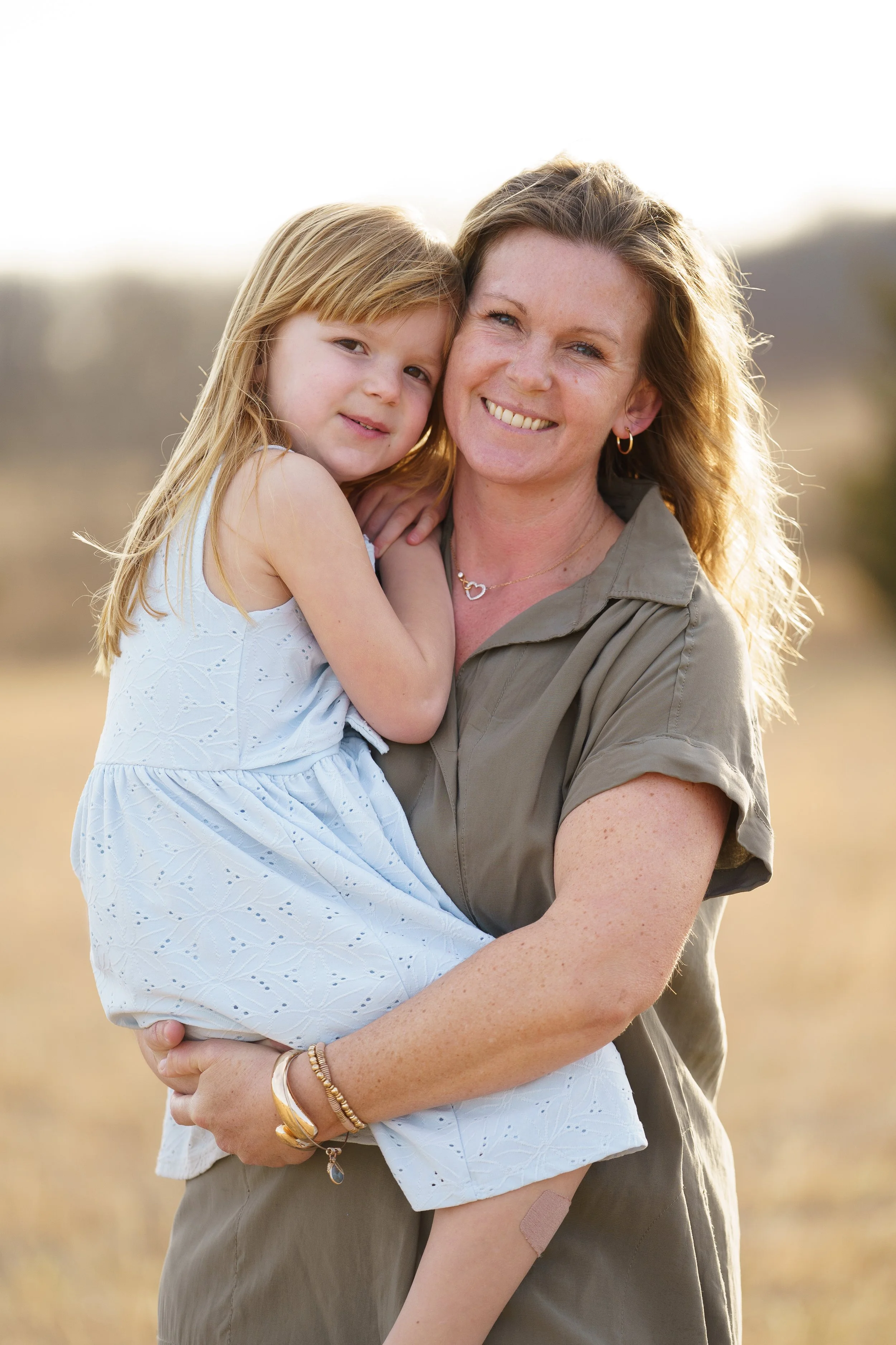 A woman with curly red hair, wearing a gray shirt, hugging a young girl with blonde hair and a white dress, standing outdoors in a field during daytime.
