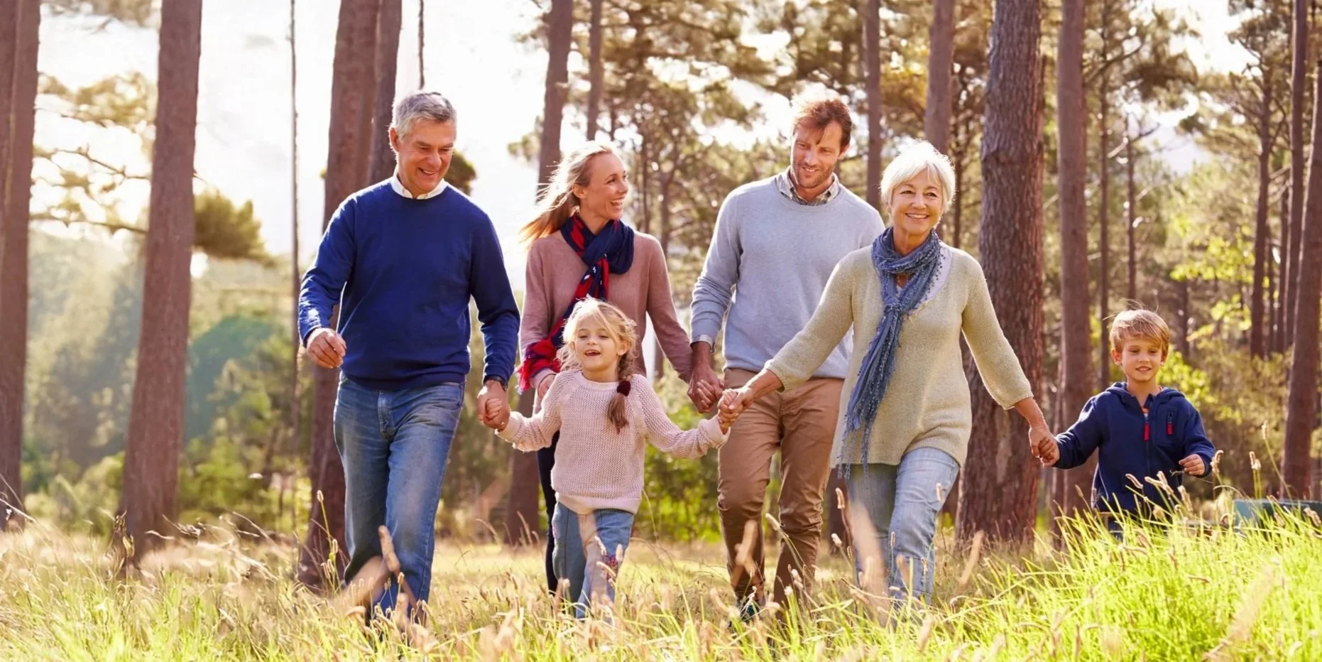 A multigenerational family enjoying a walk in a forested park on a sunny day, holding hands and smiling.