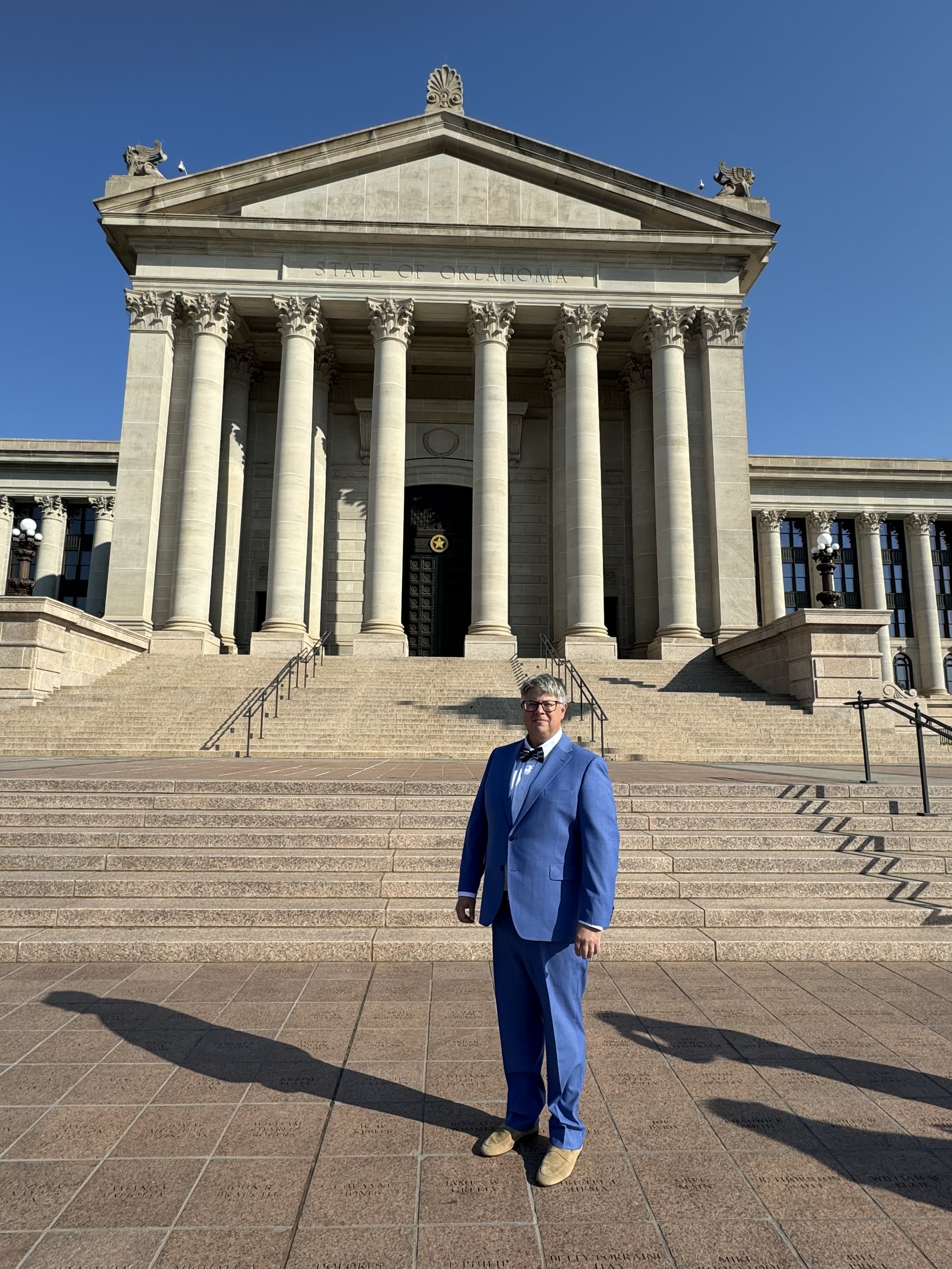 A man in a blue suit and bowtie standing on stairs in front of the Oklahoma State Capitol building, which has large columns and a triangular pediment, under a clear blue sky.