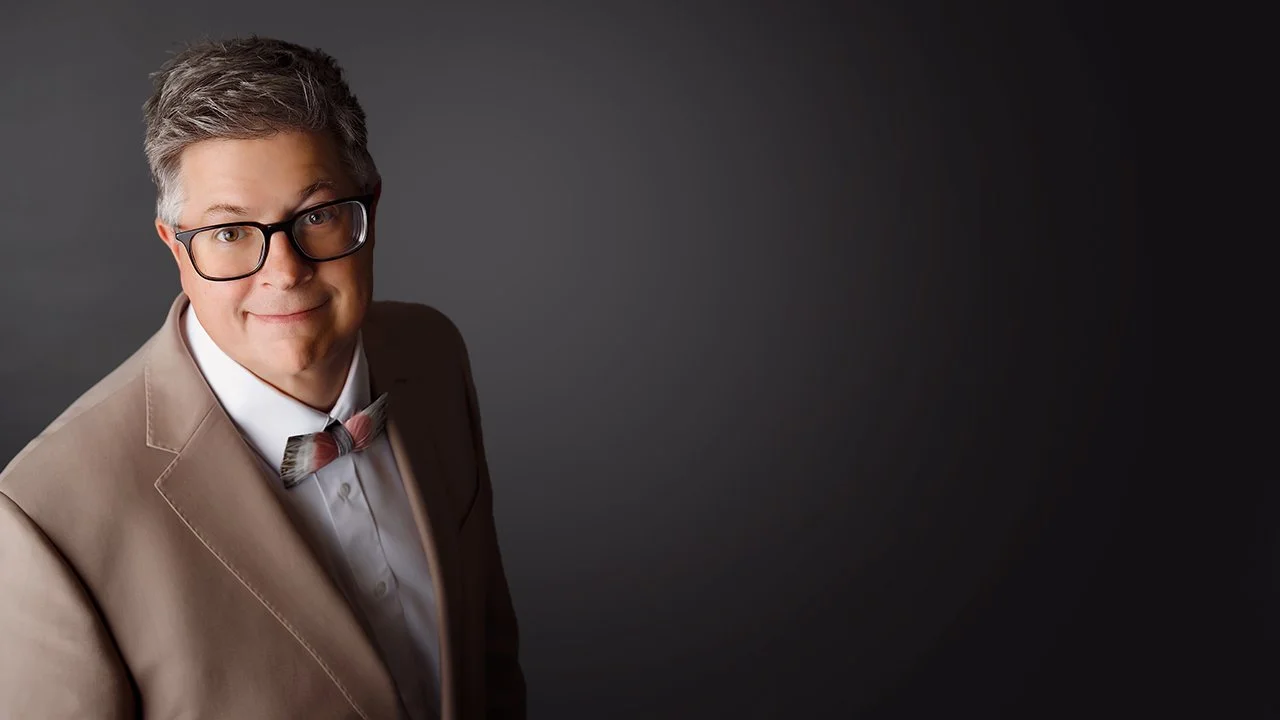 A man with gray hair, glasses, wearing a beige suit, white shirt, and bow tie, standing against a dark gray background.