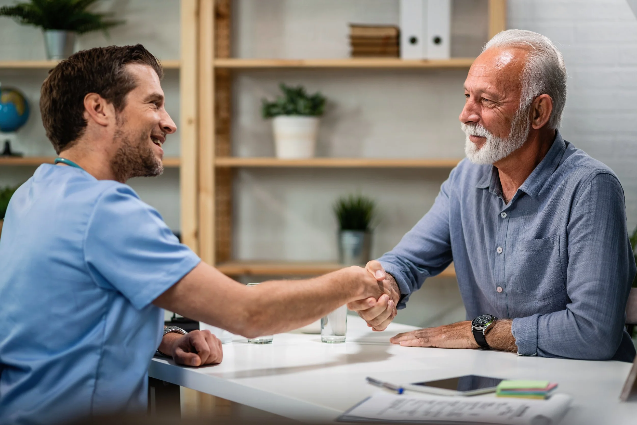 A doctor in scrubs shaking hands with an elderly man in an office setting.