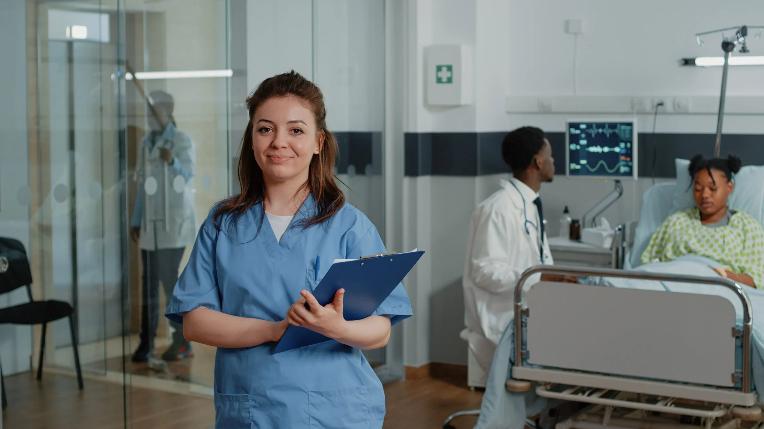 Healthcare worker in scrubs holding a clipboard, with a doctor and patient in a hospital room, medical equipment and monitors visible in the background.
