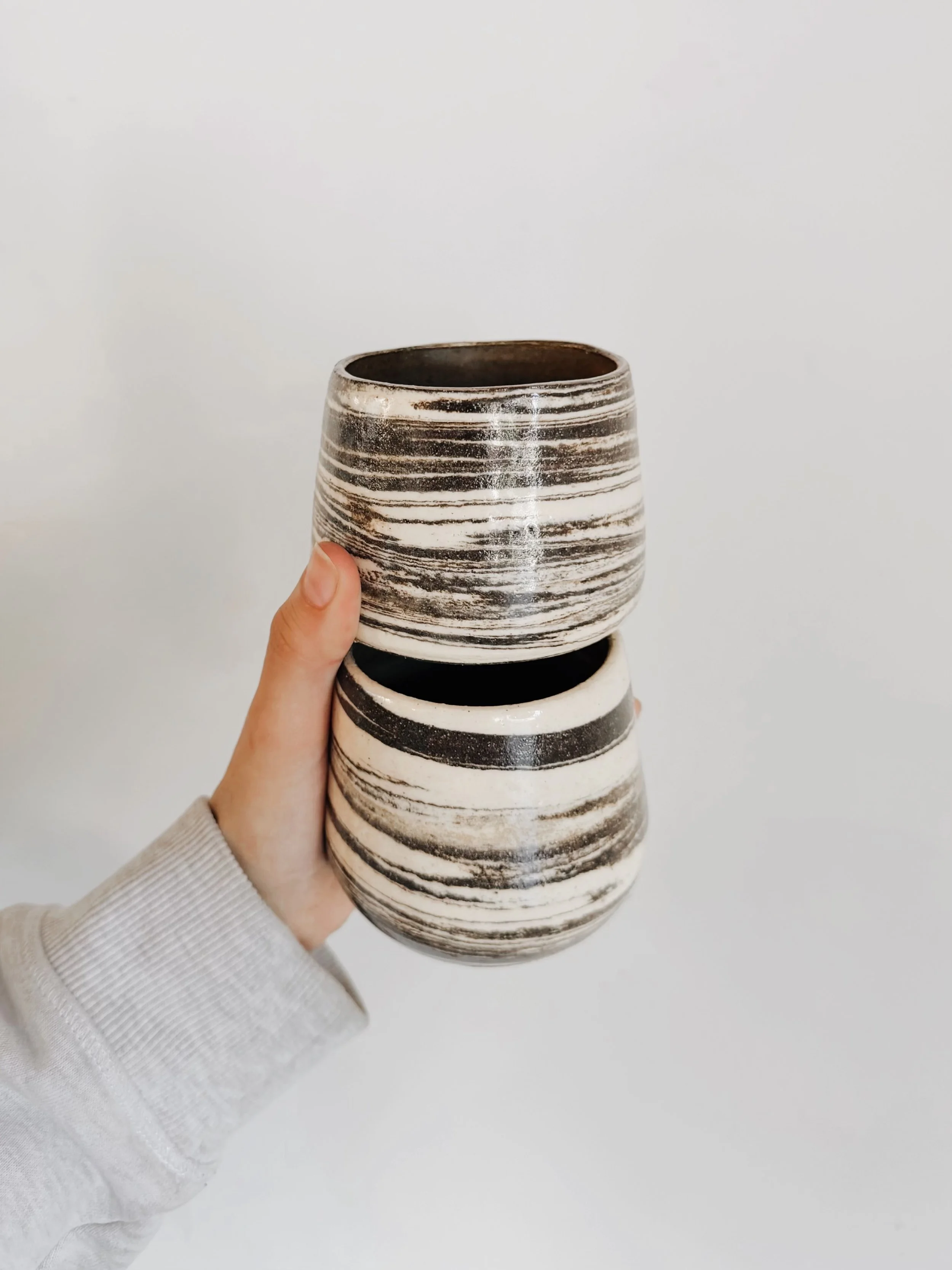 A person holding two ceramic pots with black and white horizontal striped patterns against a plain white background.