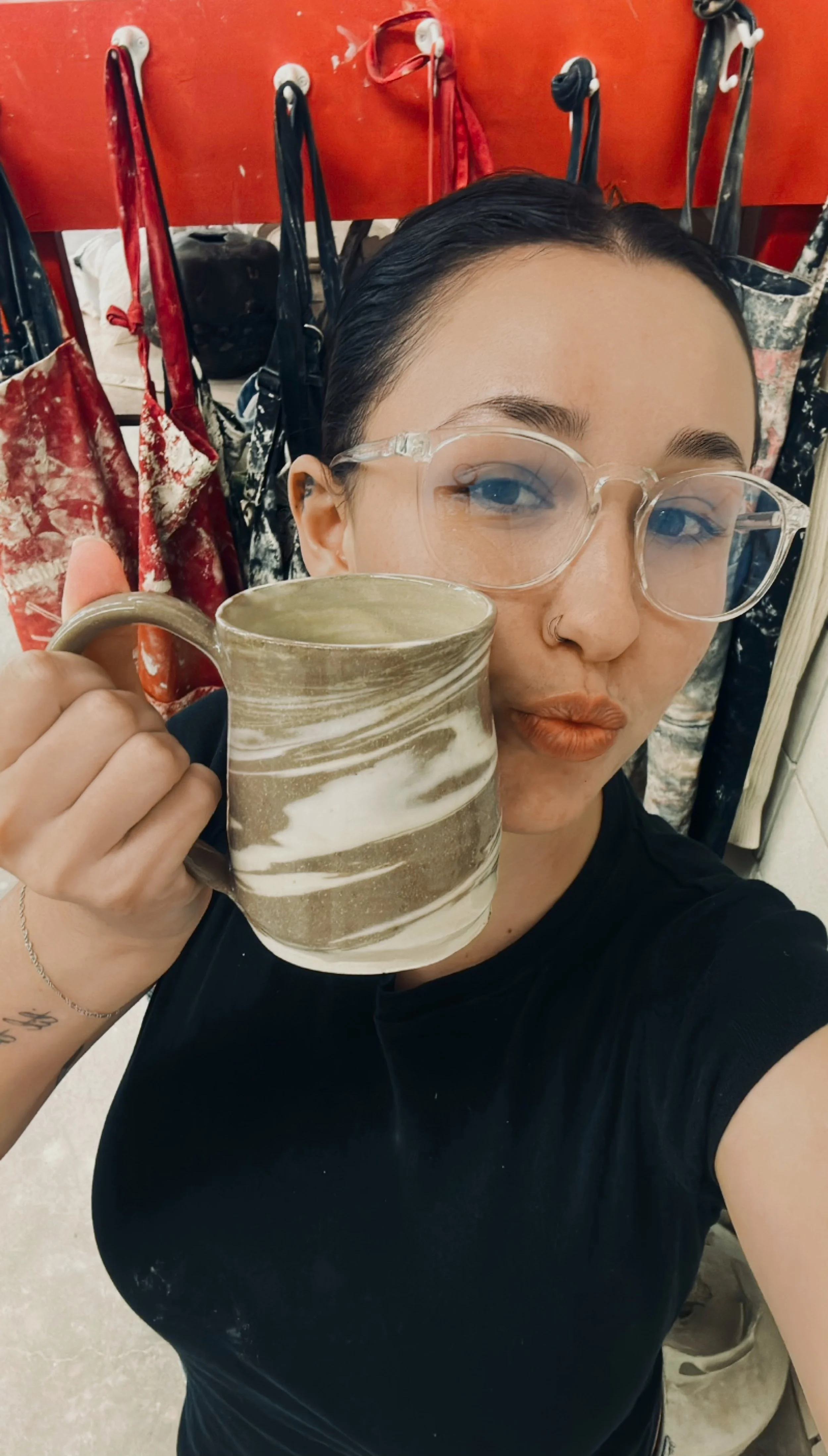 A woman with glasses and a nose ring holding a marbled ceramic mug, standing in front of a red wall with hanging aprons.