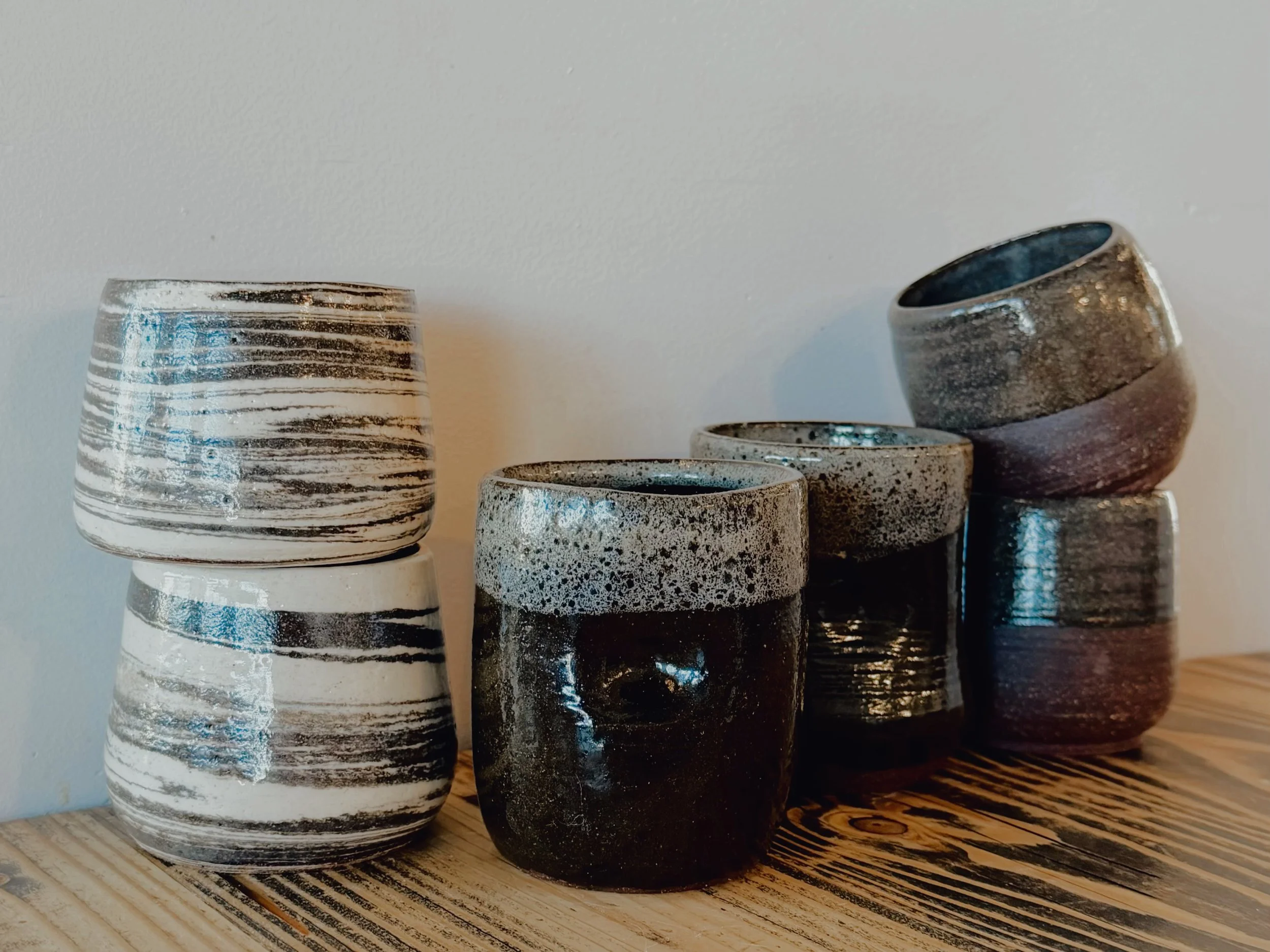 Six ceramic cups with various black, white, and gray glaze patterns, stacked in two columns on a wooden surface against a plain white wall.