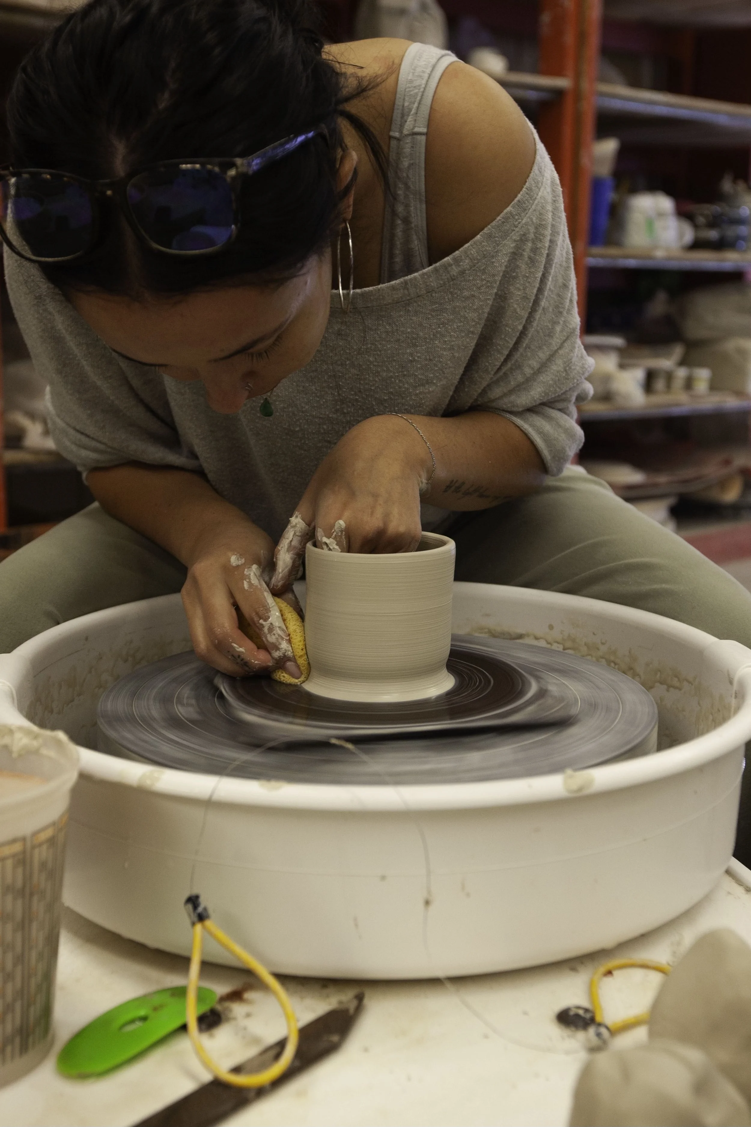 Woman shaping a ceramic pot on a pottery wheel, wearing glasses and a gray shirt, in a ceramic studio.