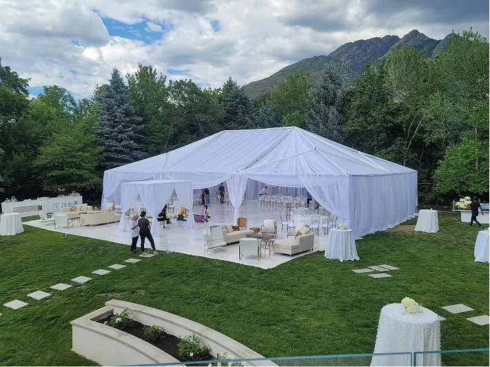 Large white event tent set up outdoors on a grassy area, with surrounding furniture and table arrangements for a celebration, with mountain scenery and trees in the background.