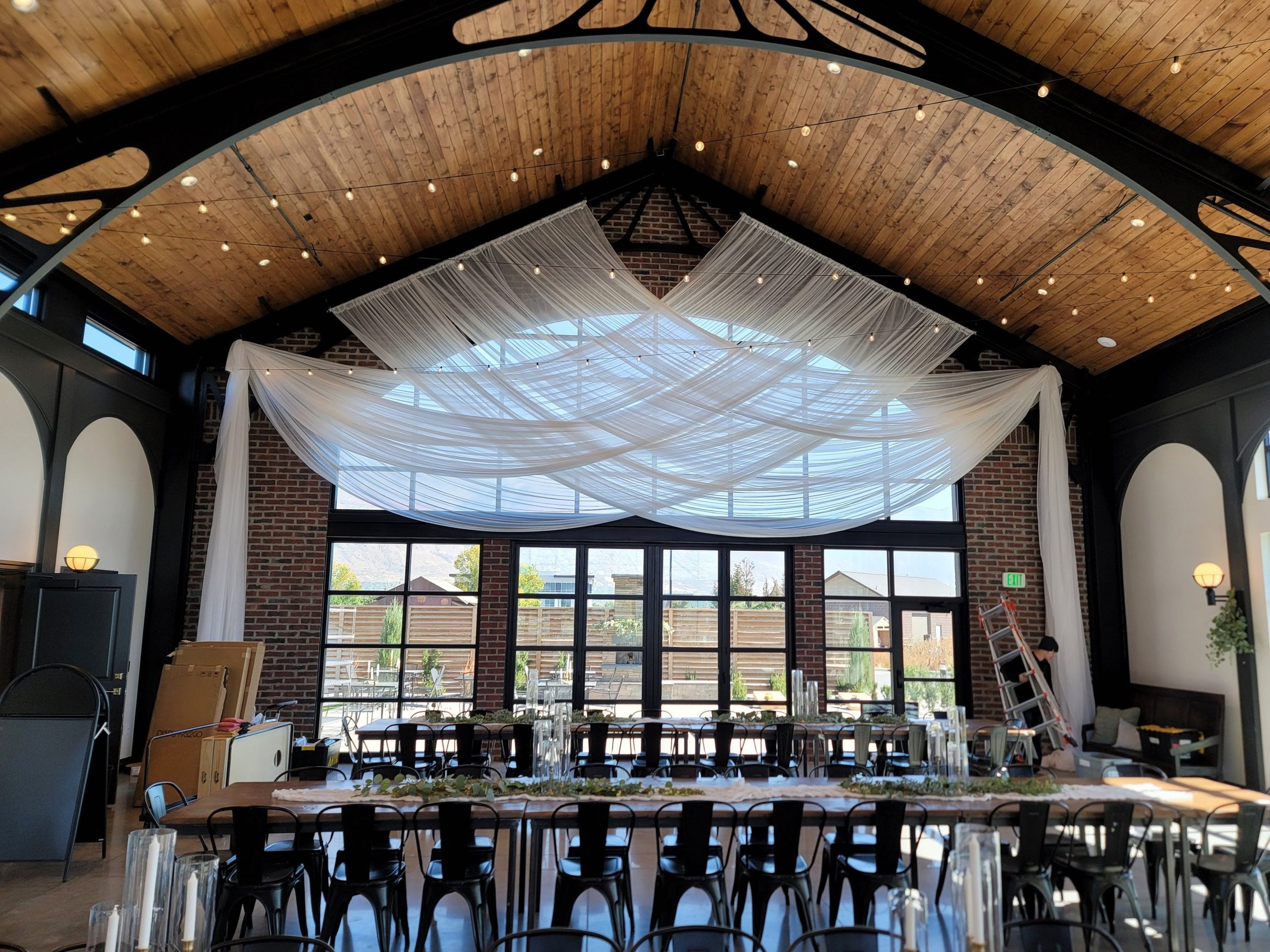 Interior of a decorated rustic event space with exposed brick walls and wood ceiling, featuring draped white fabric and hanging lights. Long tables with black chairs are arranged in front of large windows.