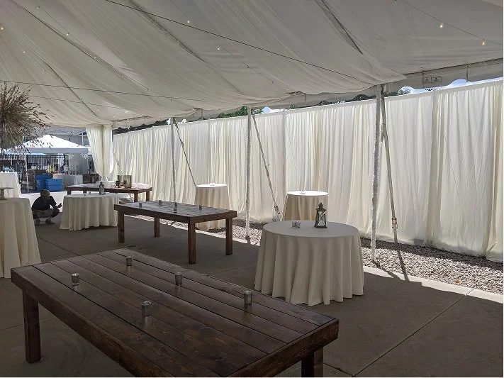 Event tent interior with round and rectangular tables, some with candles and decorations, under a white canopy with side curtains, set up for a gathering or celebration.