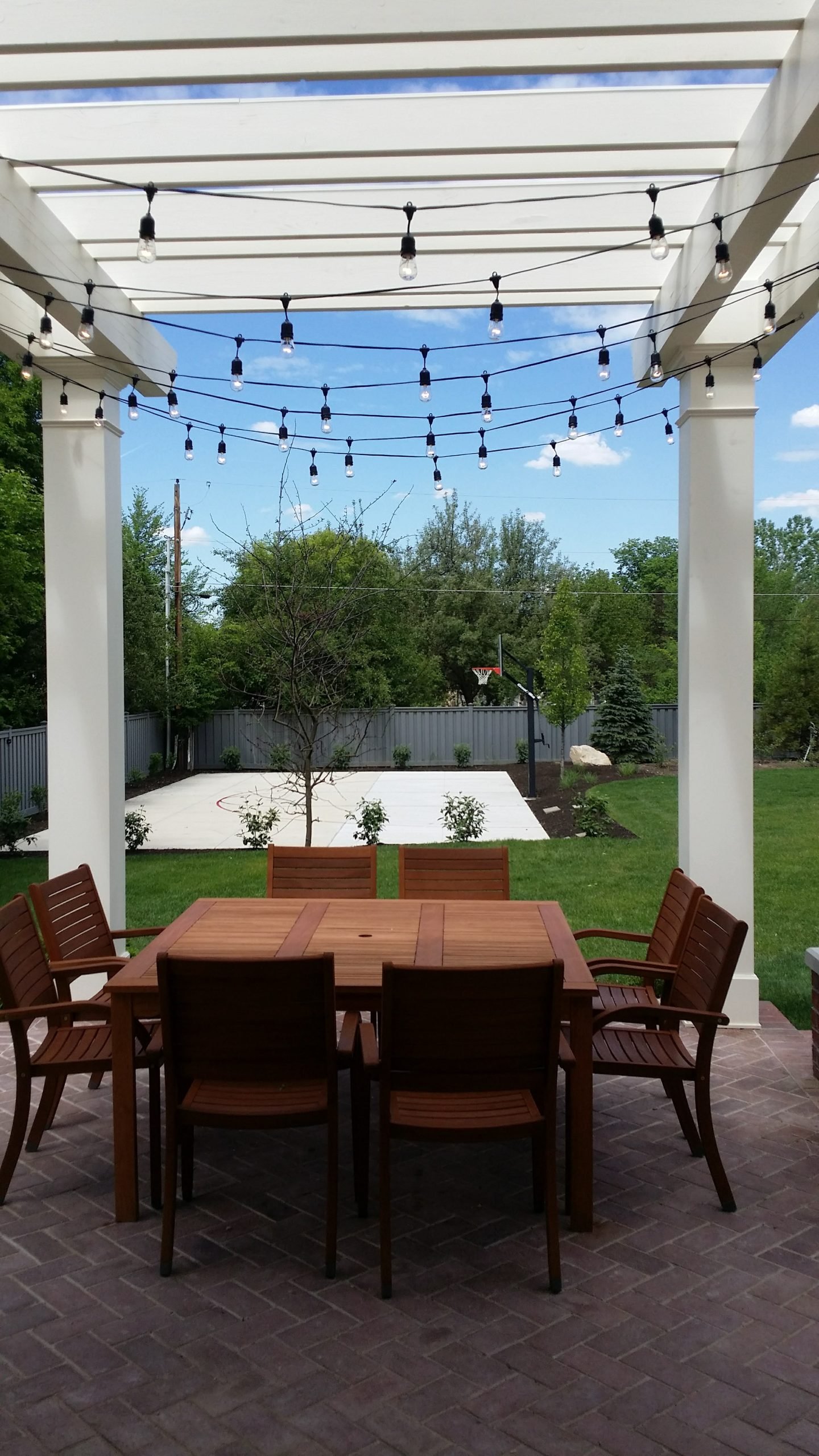 Backyard patio with pergola and string lights, featuring an outdoor wooden dining set and a view of a small basketball court.