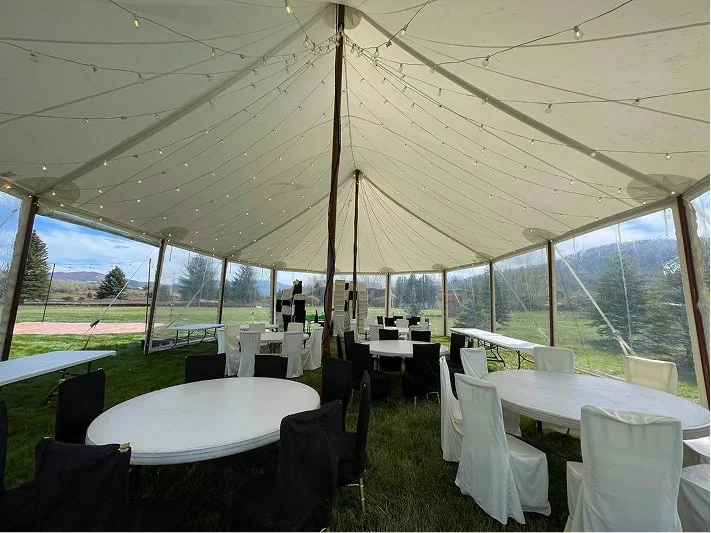 Underground event tent with string lights, round tables, and chairs in black and white, set in a grassy outdoor area with a scenic view of trees and mountains.