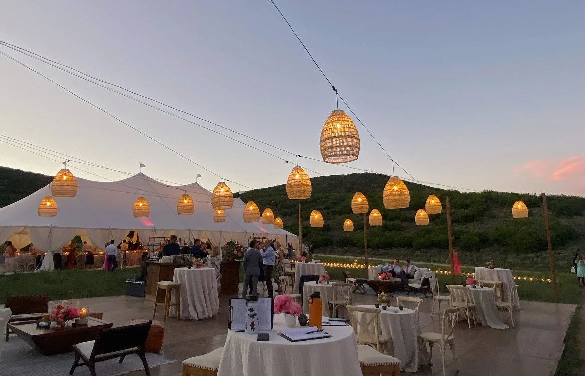 Outdoor evening event with string lights and a large white tent on a grassy hillside.