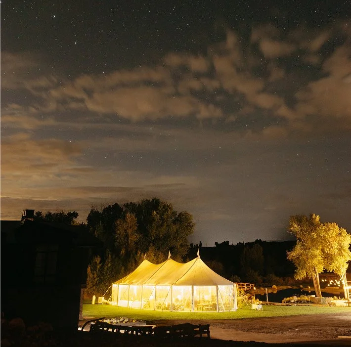 A large illuminated tent set up outdoors at night, with trees in the background and a cloudy starry sky overhead.