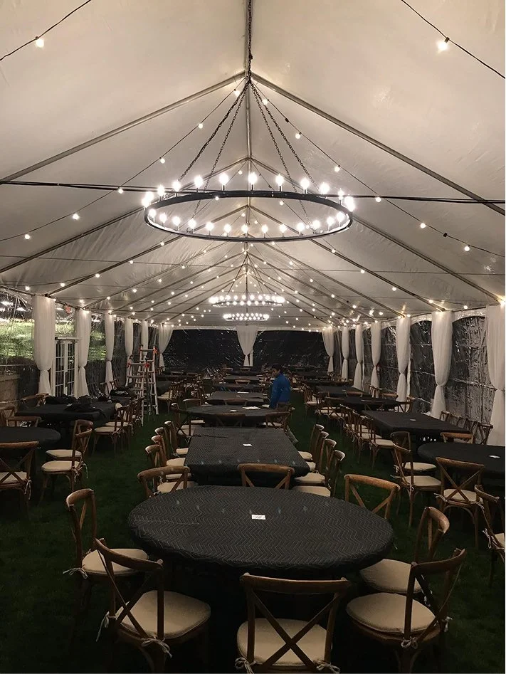 Interior of a large event tent decorated with string lights and chandeliers, set up with multiple tables and chairs for a gathering or celebration, with some staff adjusting the arrangements.