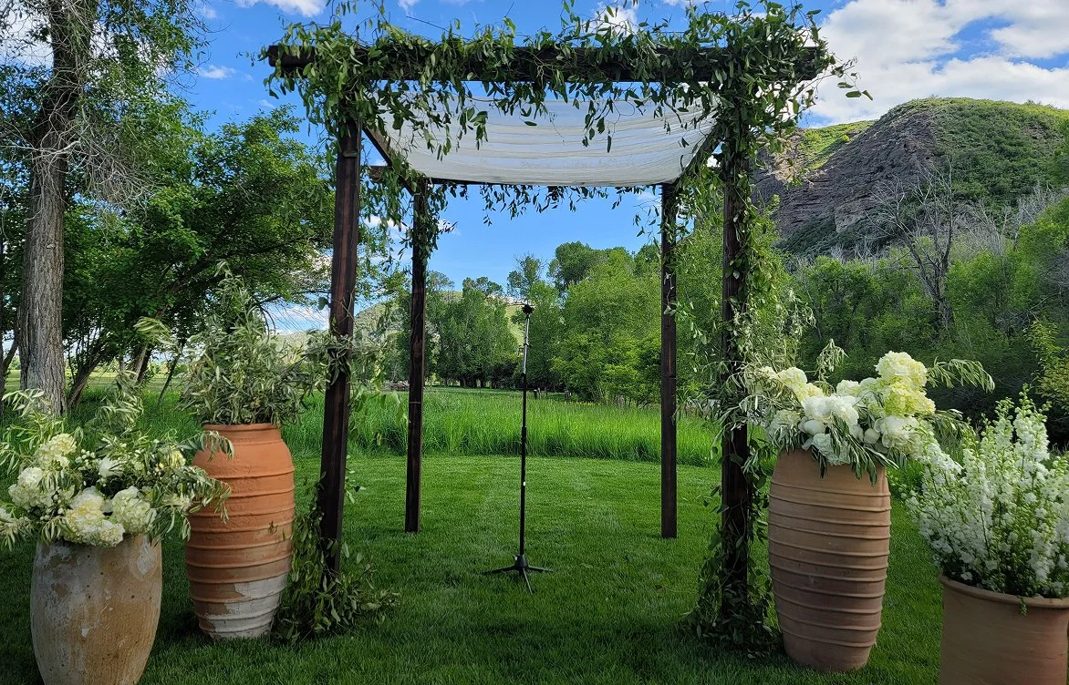An outdoor wedding setup with a wooden arbor draped in green leaves and white fabric, flanked by large white pots filled with white flowers on a grassy lawn with trees, hills, and a blue sky in the background.