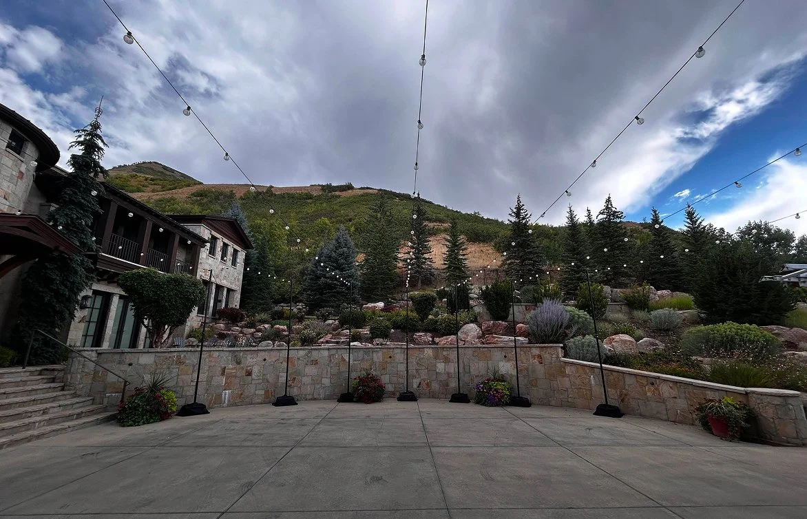 Outdoor patio with string lights, stone wall, potted plants, and a hillside with trees and mountains in the background.