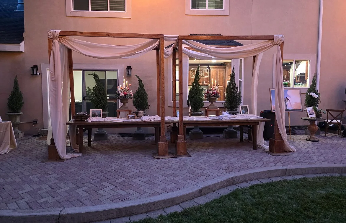 Outdoor wedding or event setup with a wooden canopy and draped fabric, tables with candles and floral arrangements, potted trees, and framed photographs, set on a brick patio at dusk.