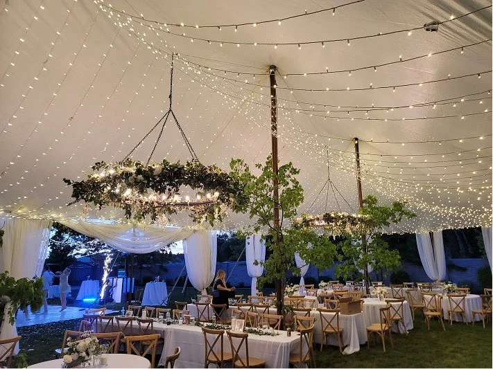A decorated outdoor event tent with tables and chairs, hanging floral arrangements, string lights, and draped fabric, set for a celebration or wedding reception.