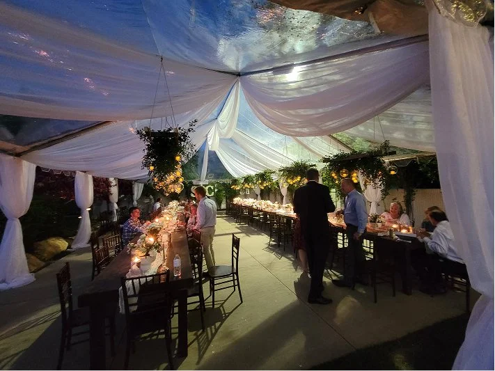 A daytime outdoor dining event under a white tent with draped fabric, decorated with hanging greenery and yellow flowers, featuring long tables with lit candles, chairs, and people mingling and eating.