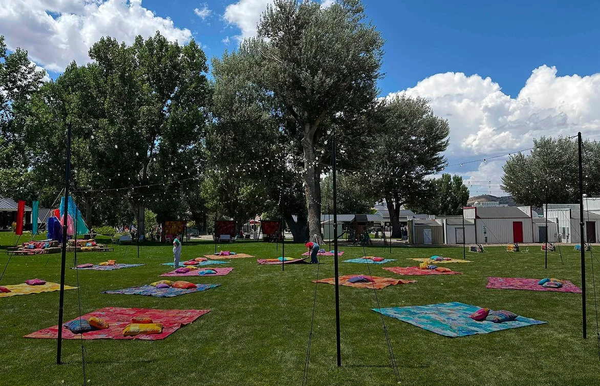 Outdoor scene with colorful mats and pillows on grass, string lights hanging, large trees, and small white buildings in the background under partly cloudy sky.