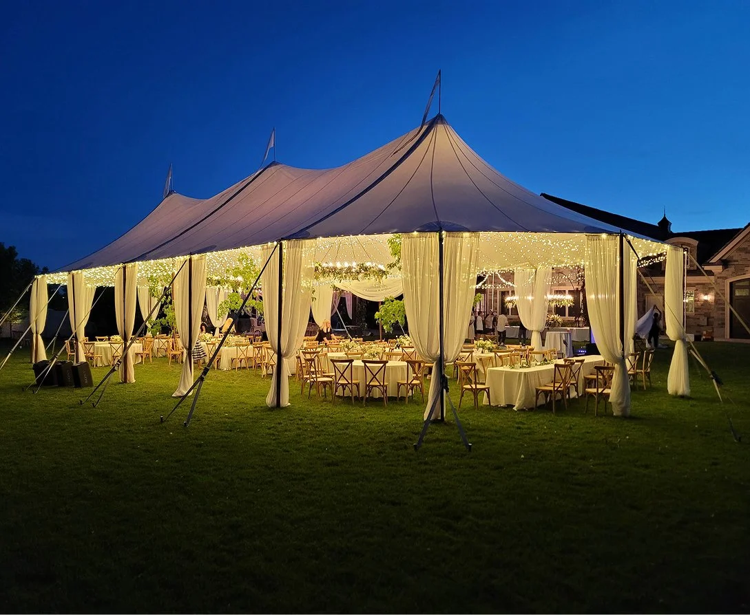 Outdoor wedding reception under a large illuminated tent at dusk with decorated tables and chairs on a grassy lawn.