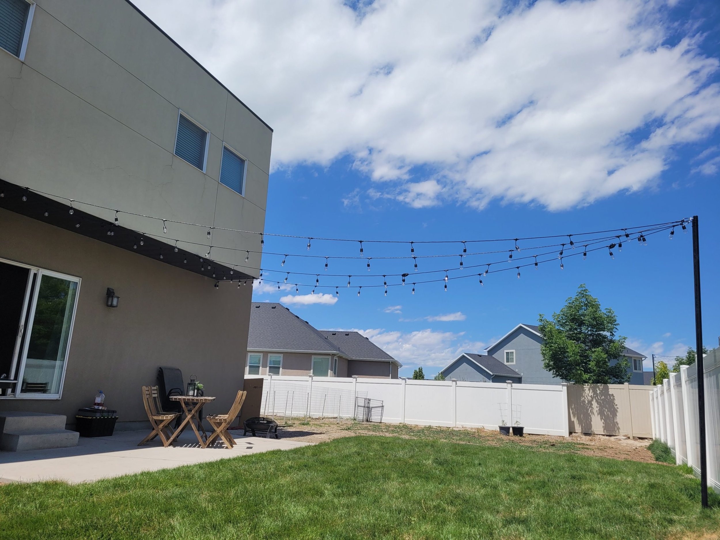 A backyard patio area with string lights, a small wooden table and chairs, a beige house, white fence, and neighboring houses under a blue sky with clouds.