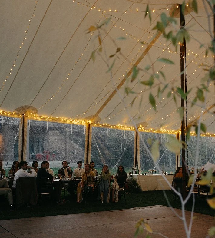 People seated at a long banquet table inside a decorated tent with string lights and greenery, during an outdoor event or celebration.