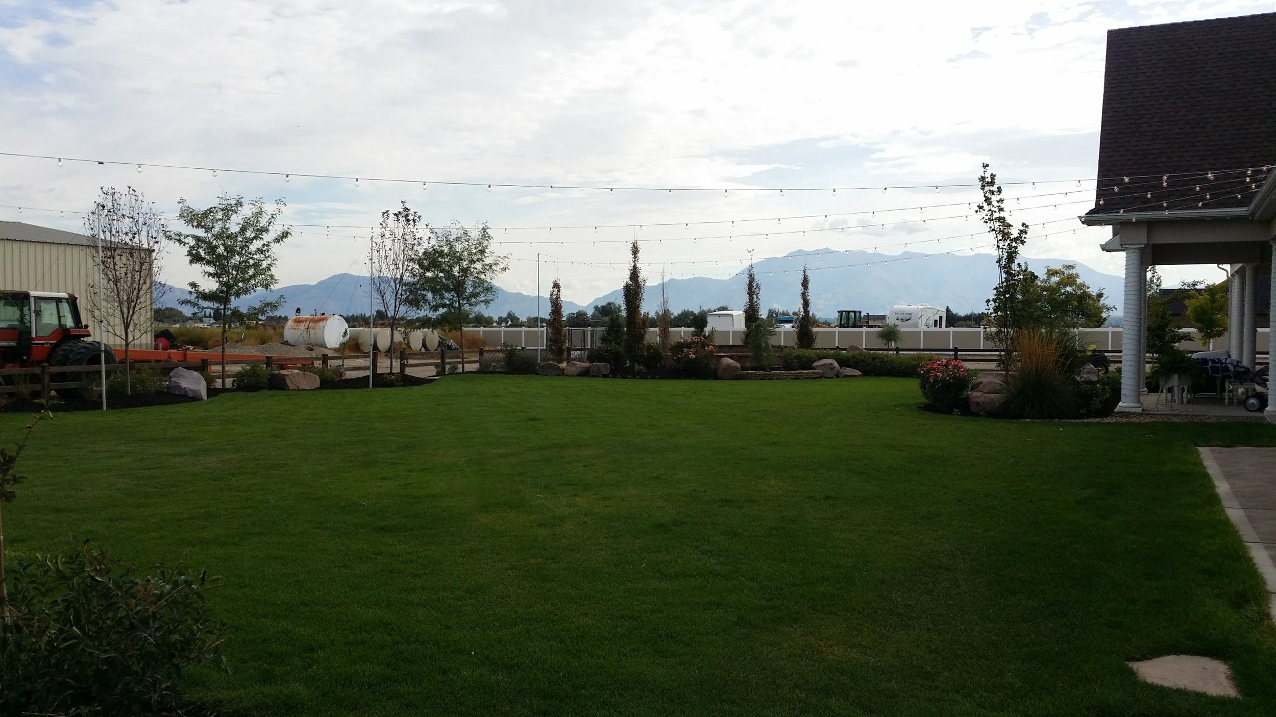 A spacious green lawn is in the foreground, surrounded by a garden with small trees and shrubs. String lights are hung above, and a tractor is visible on the left next to some tanks. In the background, mountains are visible under a cloudy sky. Part o
