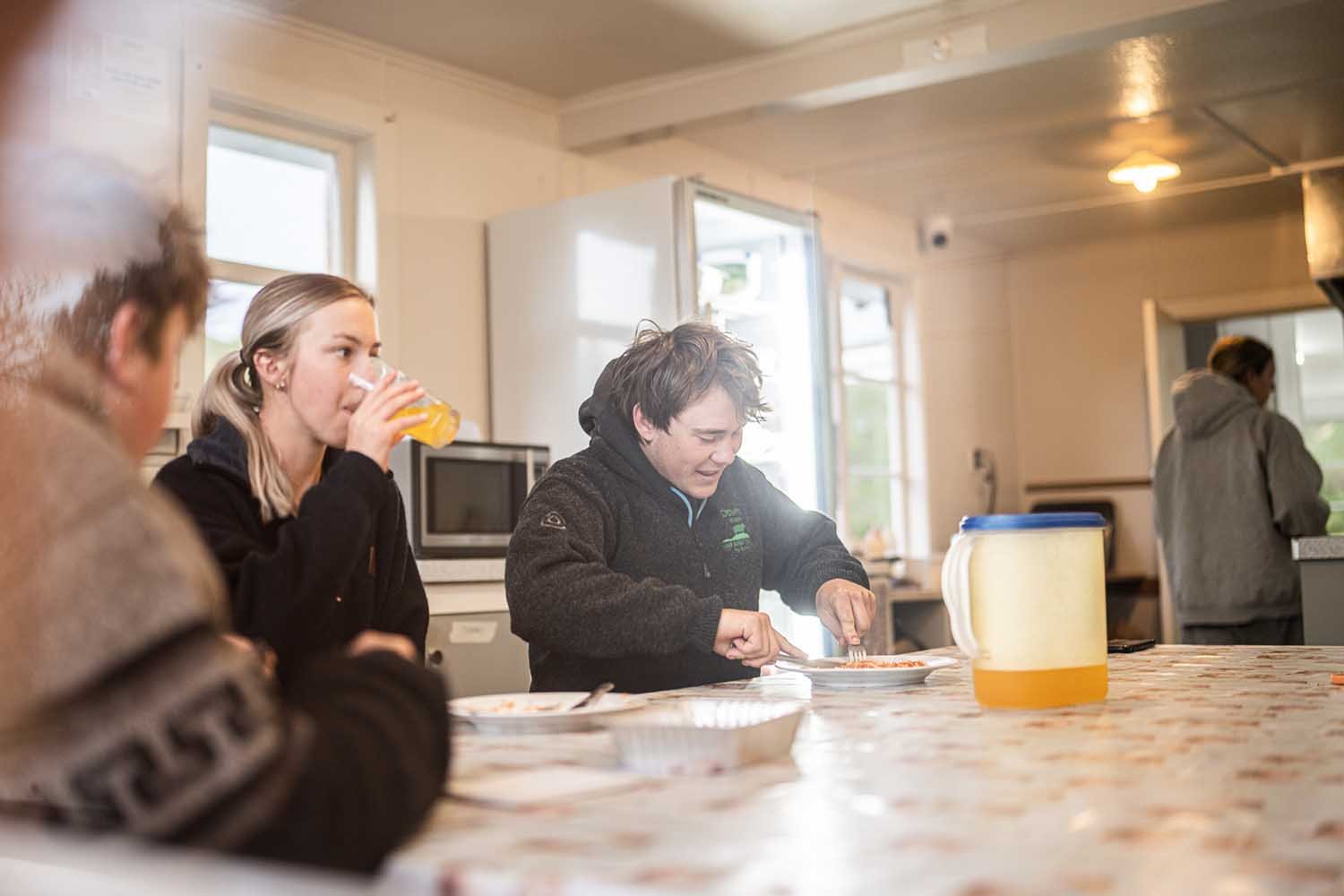 People sitting at a kitchen table, one drinking juice, another eating, with a jug on the table.