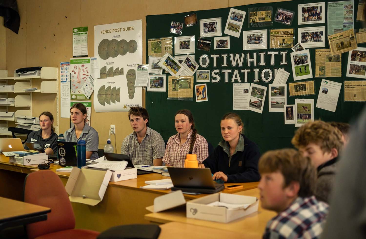 A group of young people sit at desks in a classroom setting. They are focused and attentive, with laptops and notebooks in front of them. The wall behind them is covered with posters and photographs, including one that reads 'Otiwhiti Station'. The e