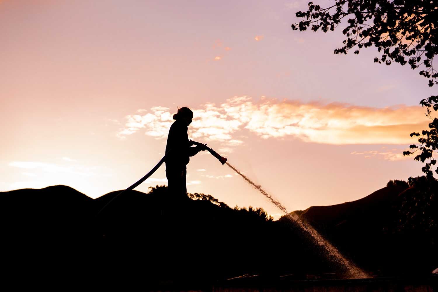 Silhouette of a firefighter spraying water with a hose at sunset, with a scenic background of trees and hills.