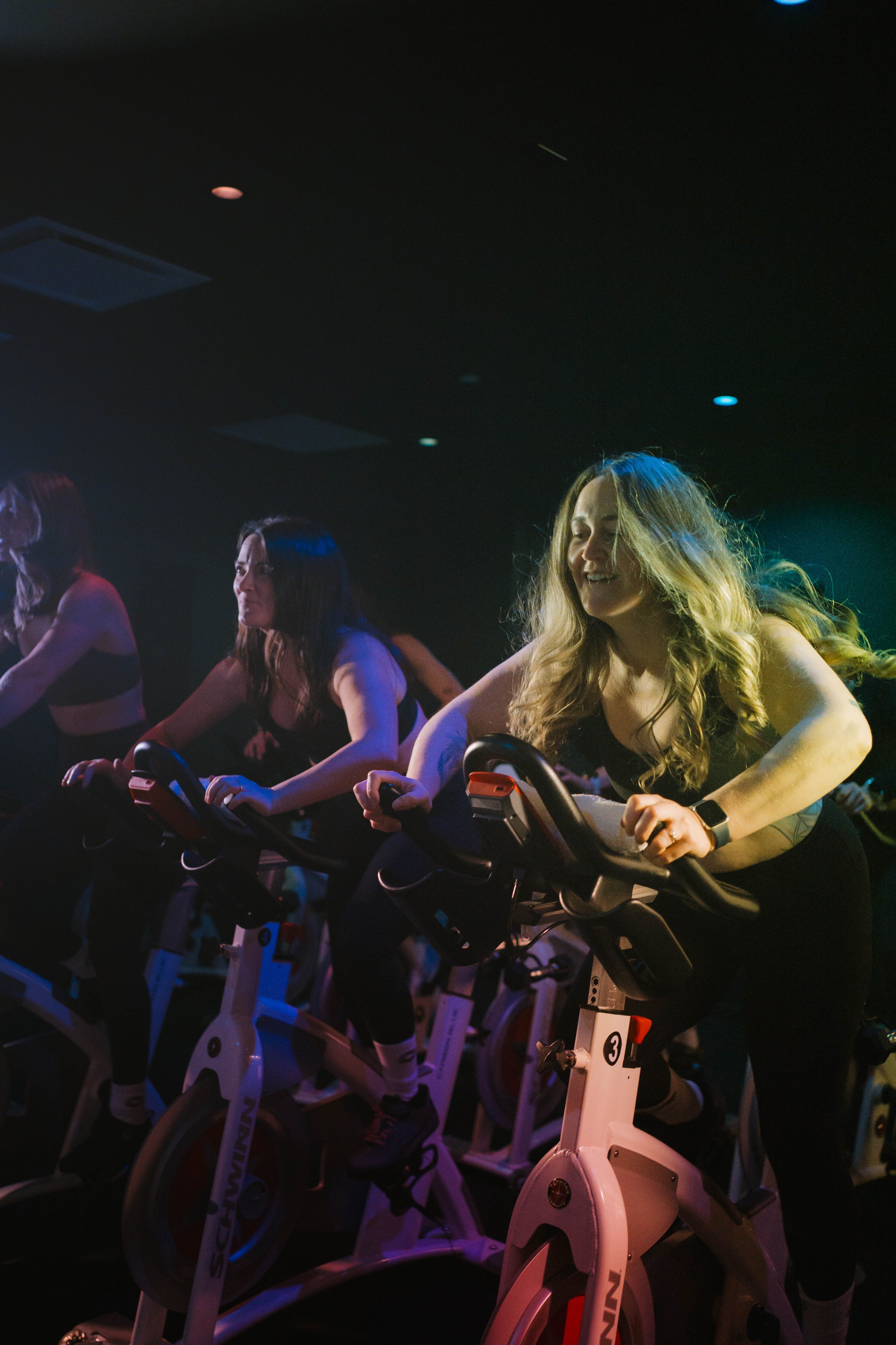 Women participating in a group indoor cycling class in a dark room with blue and purple lighting, riding stationary bikes.