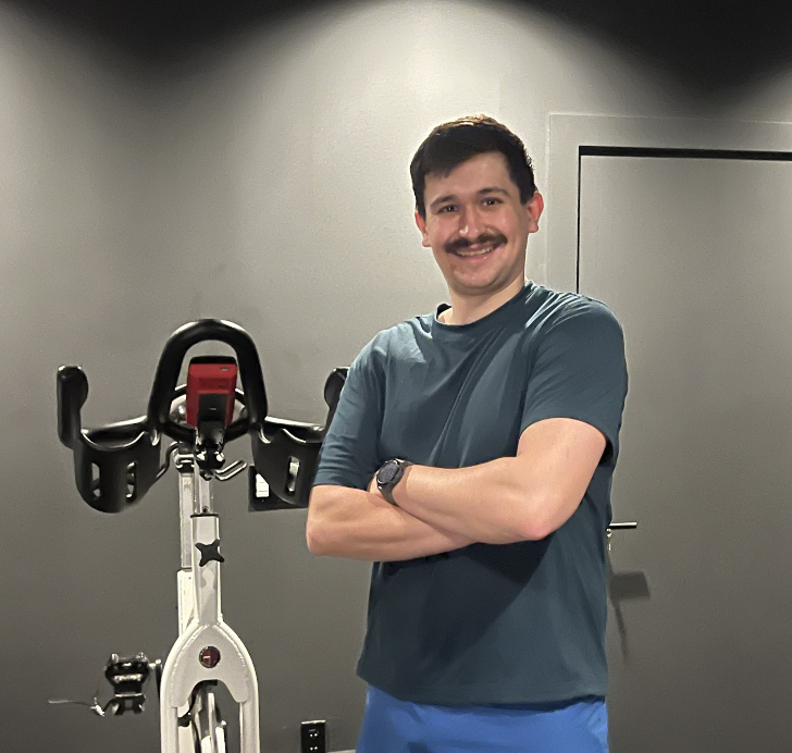 A man with a mustache, wearing a blue t-shirt and workout shorts, standing with arms crossed next to a stationary exercise bike in a gym.
