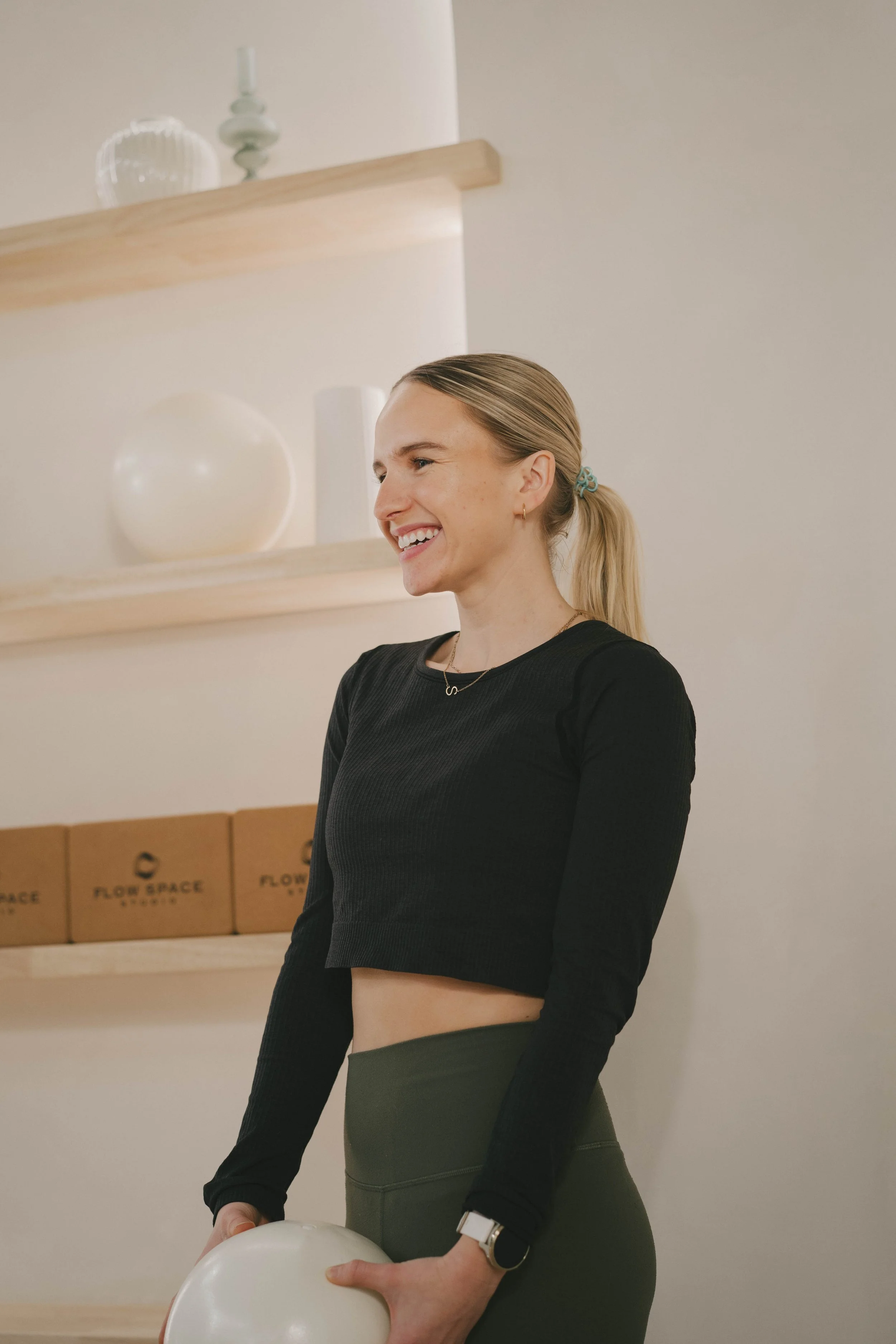 A smiling woman with blonde hair tied in a ponytail wearing a black long-sleeve shirt and green pants, holding a white balloon, standing in a room with wooden shelves and cardboard boxes in the background.