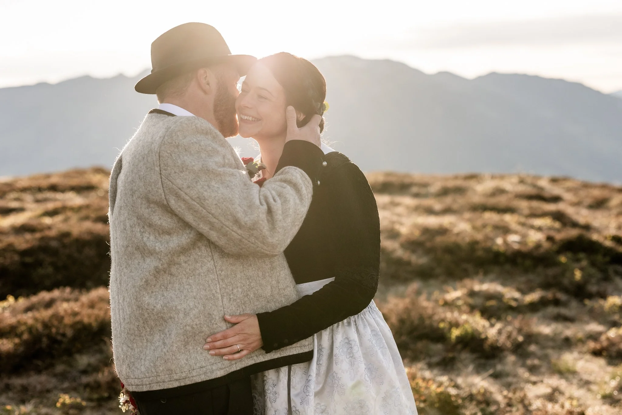 Mann führt mit seiner rechten Hand seine Frau an sich heran, flüstert ihr liebevoll ins Ohr. Das goldene Licht bei Sonnenaufgang taucht die beiden in ein warmes Licht für perfekte Hochzeitsfotos am Berg. 
