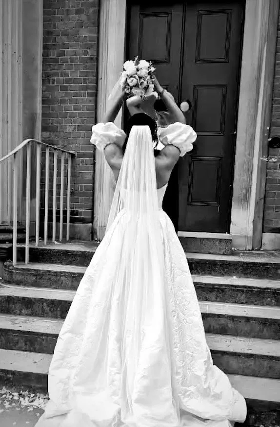 Black and white photo of a stunning white wedding gown by Wed2B, featuring a long tulle veil. The model holds a bouquet above her head, showcasing the gown’s elegant and timeless design.