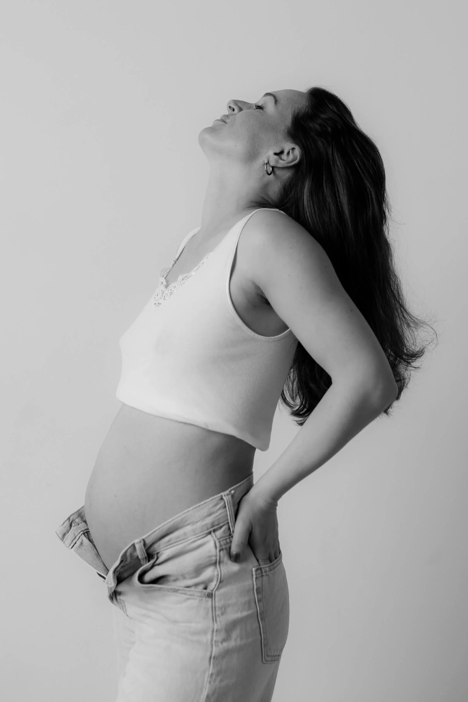 A woman with long dark hair, wearing a white sleeveless top and jeans, standing sideways with her hand in her pocket and her head tilted back with a joyful expression.