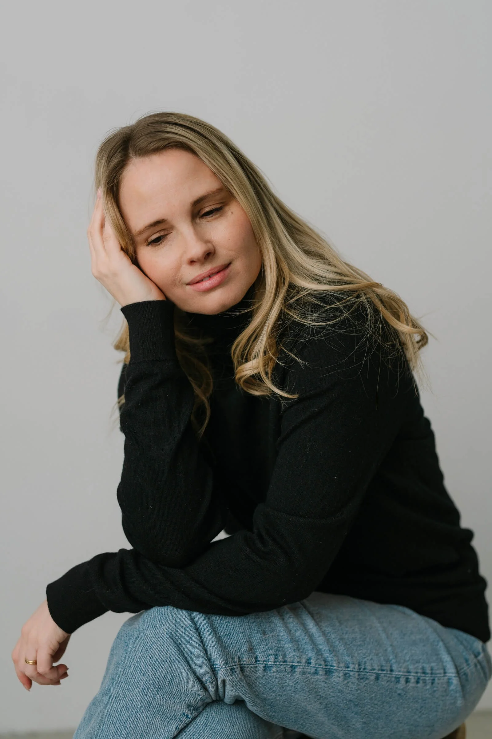 A woman with long blond hair, wearing a black sweater and light blue jeans, is sitting with her eyes closed and a pained or contemplative expression, resting her head on her left hand.