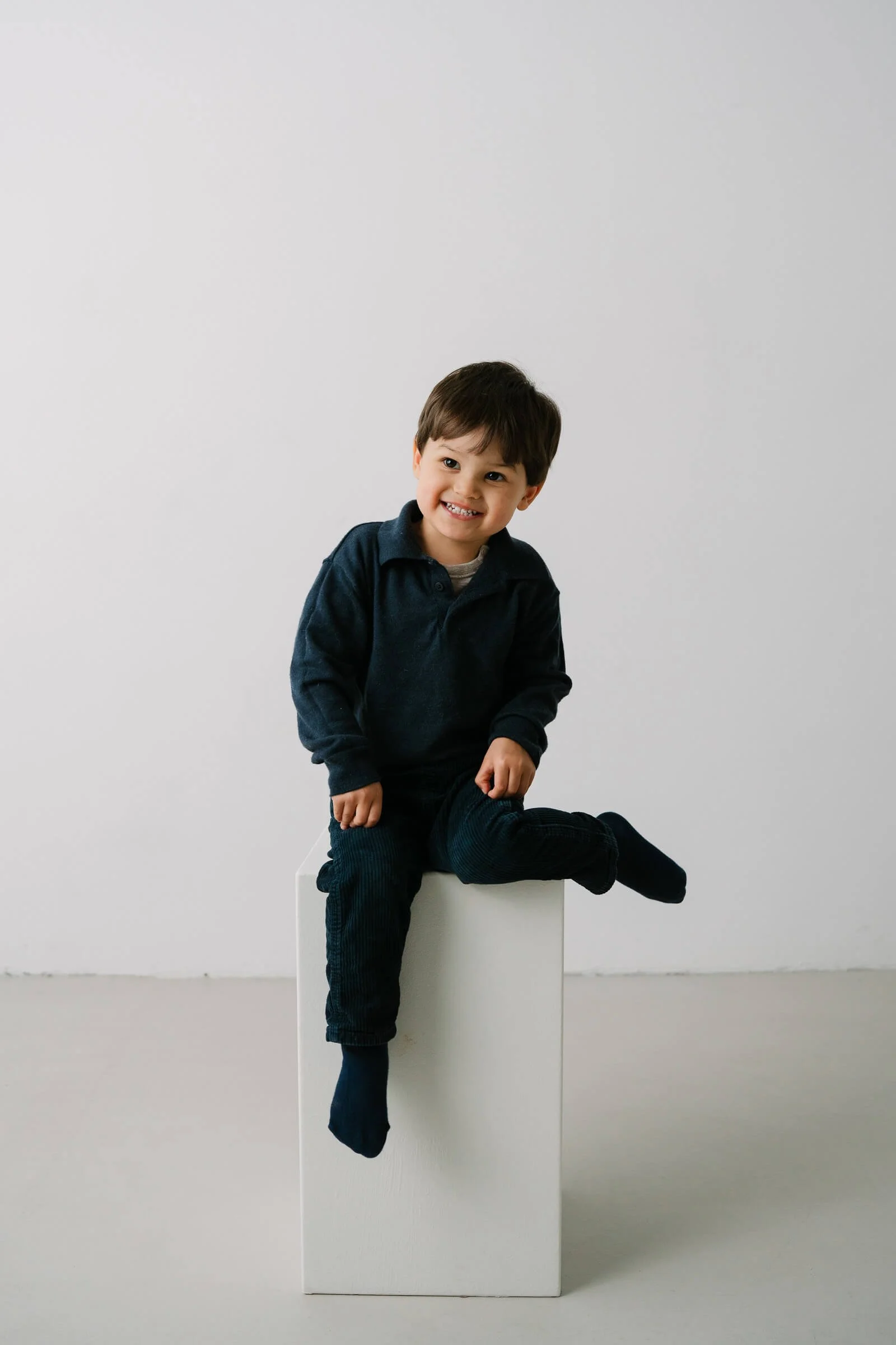 Smiling young boy sitting on a white cube in a minimalistic setting.