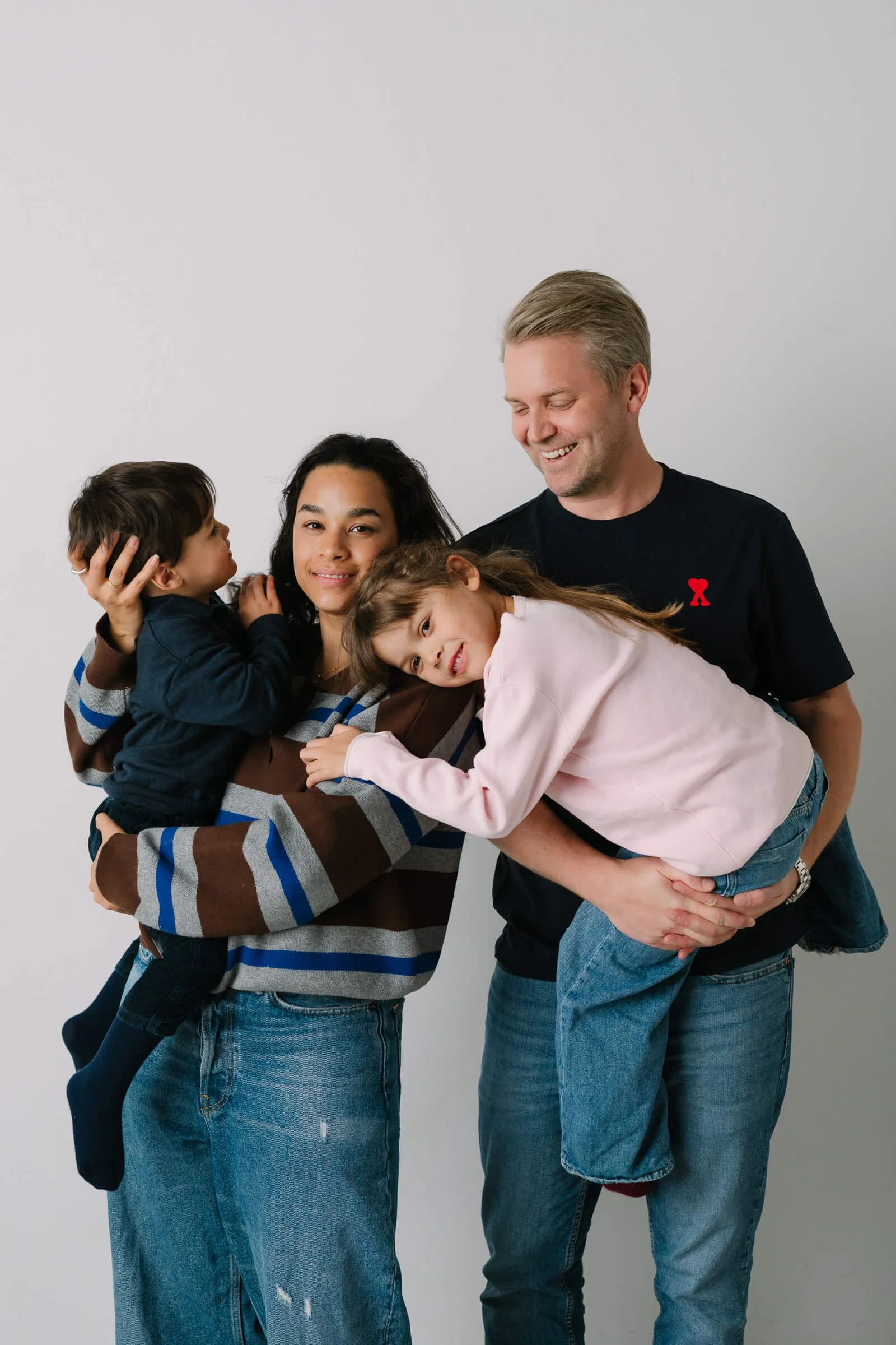 Family of four, two children and two adults, smiling and cuddling against a plain white background.
