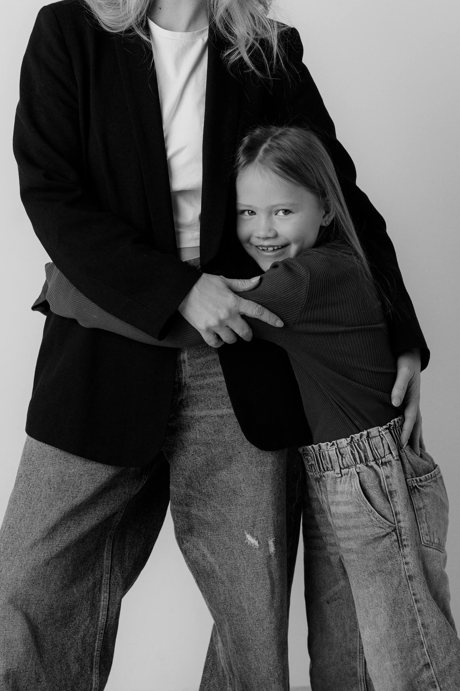 A woman and a young girl hugging each other, the woman is wearing a blazer and jeans, and the girl is wearing a long sleeve shirt and jeans.