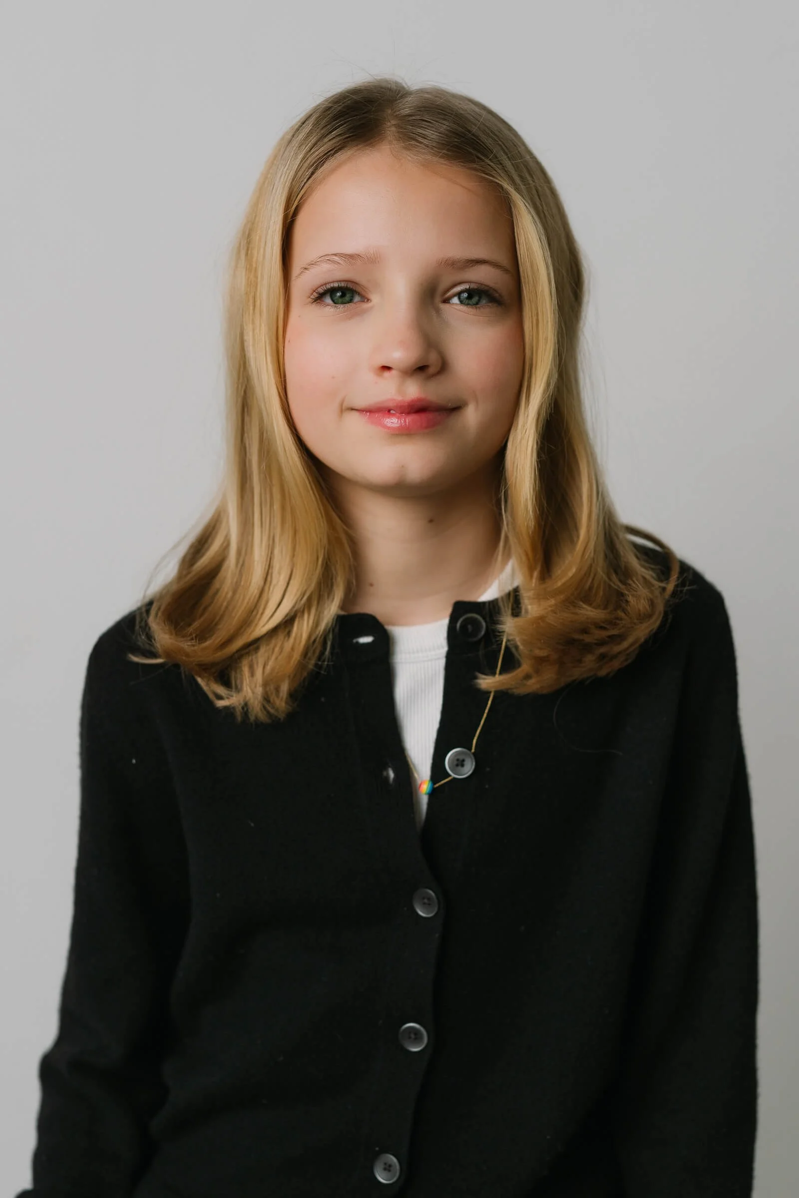 A young girl with blonde, shoulder-length hair, wearing a black cardigan and a white top, smiling slightly at the camera against a plain gray background.