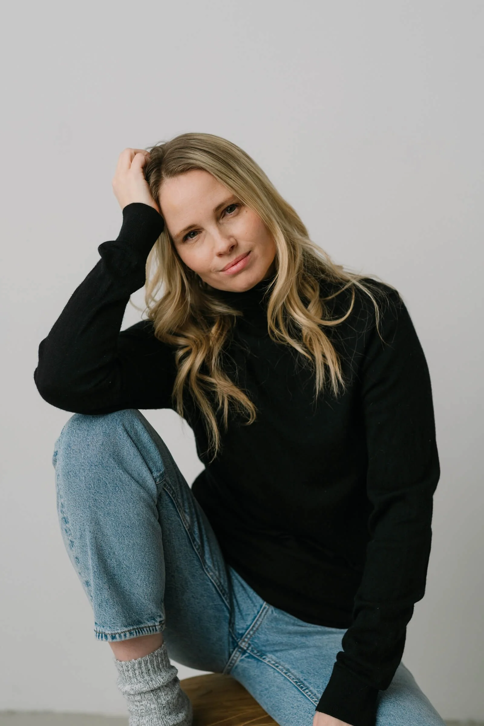 A woman with blonde wavy hair, wearing a black turtleneck and blue jeans, sitting on a wooden stool against a plain light gray background, looking at the camera with a slight smile.