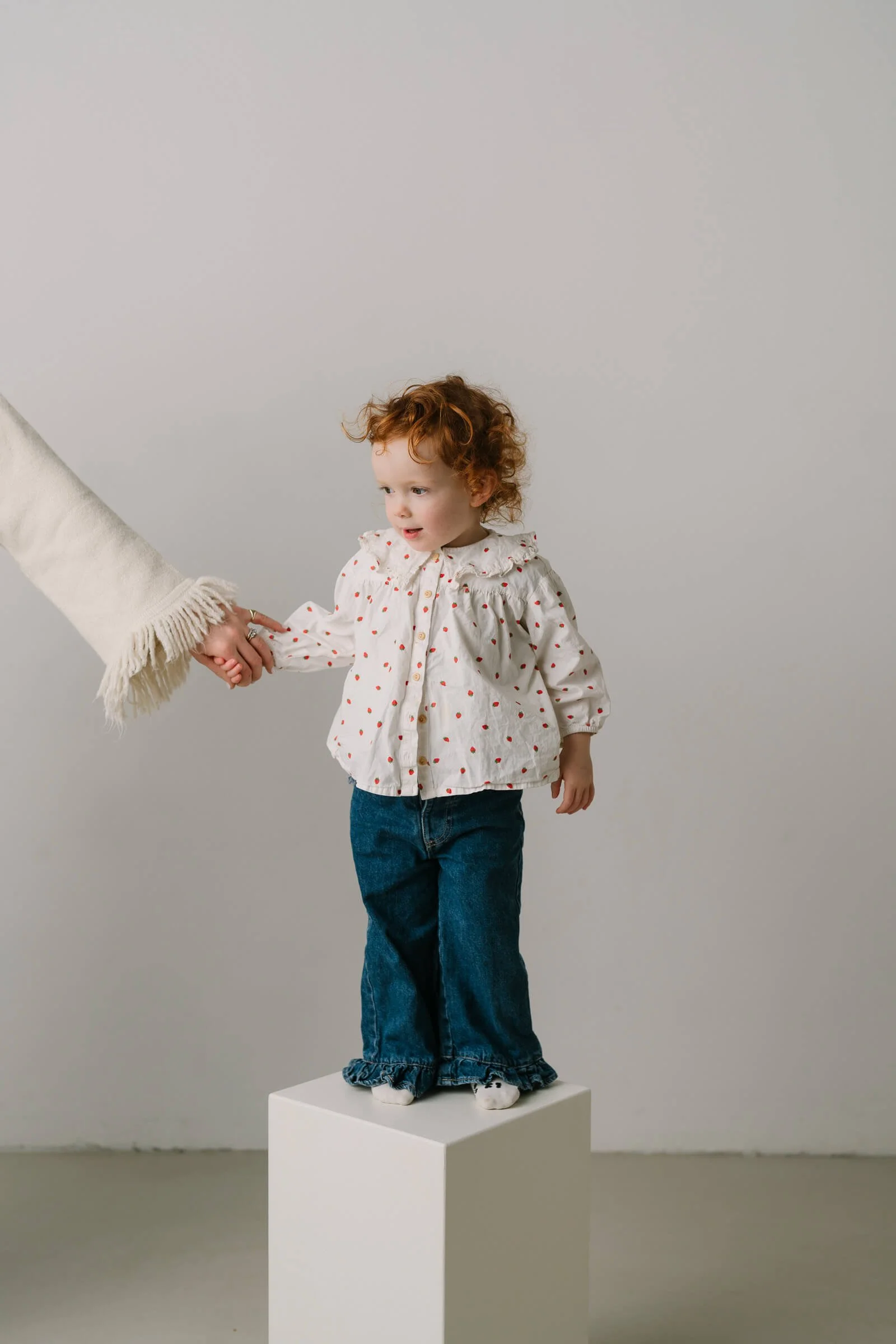 A young girl with curly red hair standing on a white pedestal, holding hands with an adult.