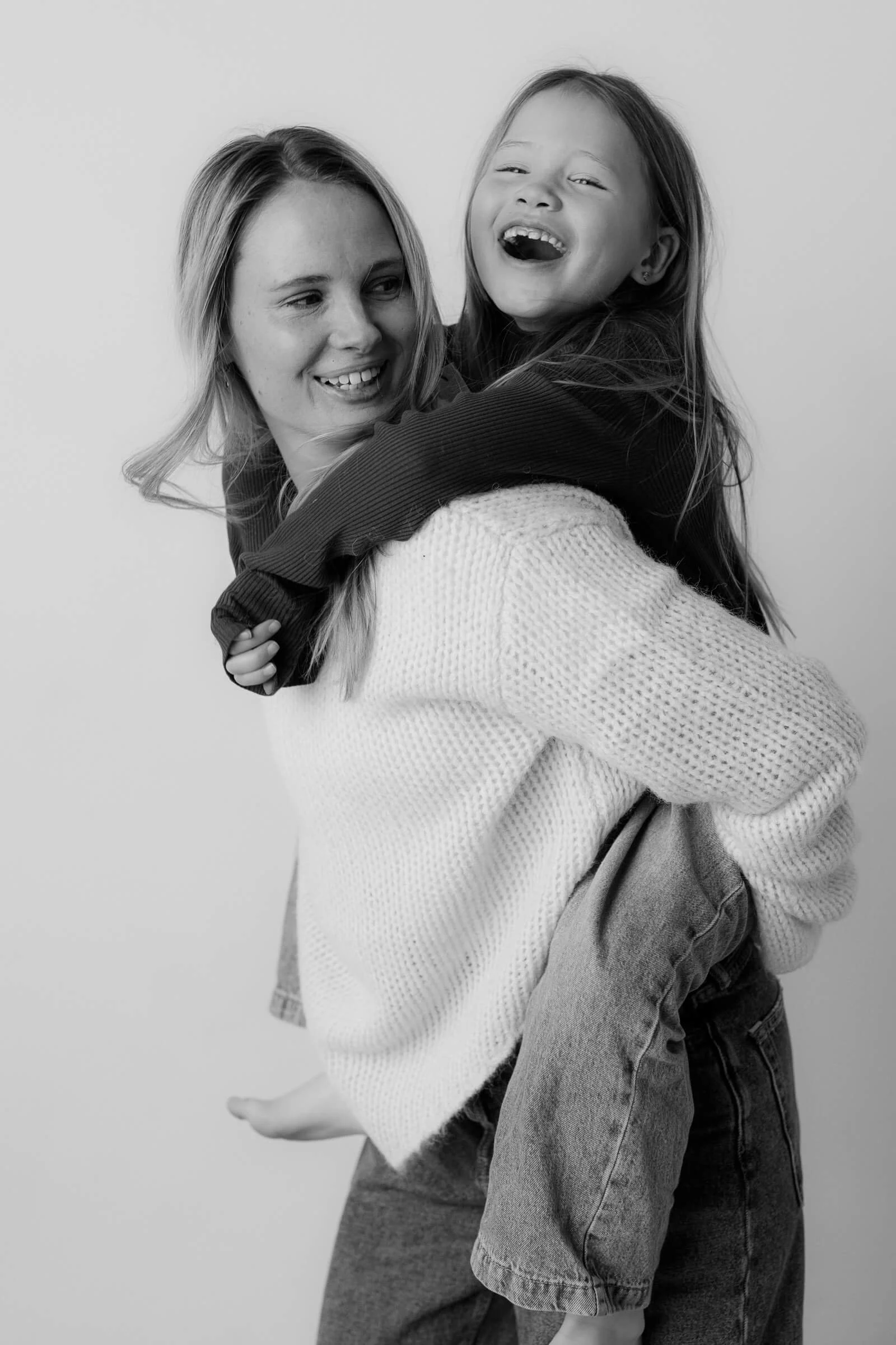 A woman giving a piggyback ride to a laughing young girl against a plain background.