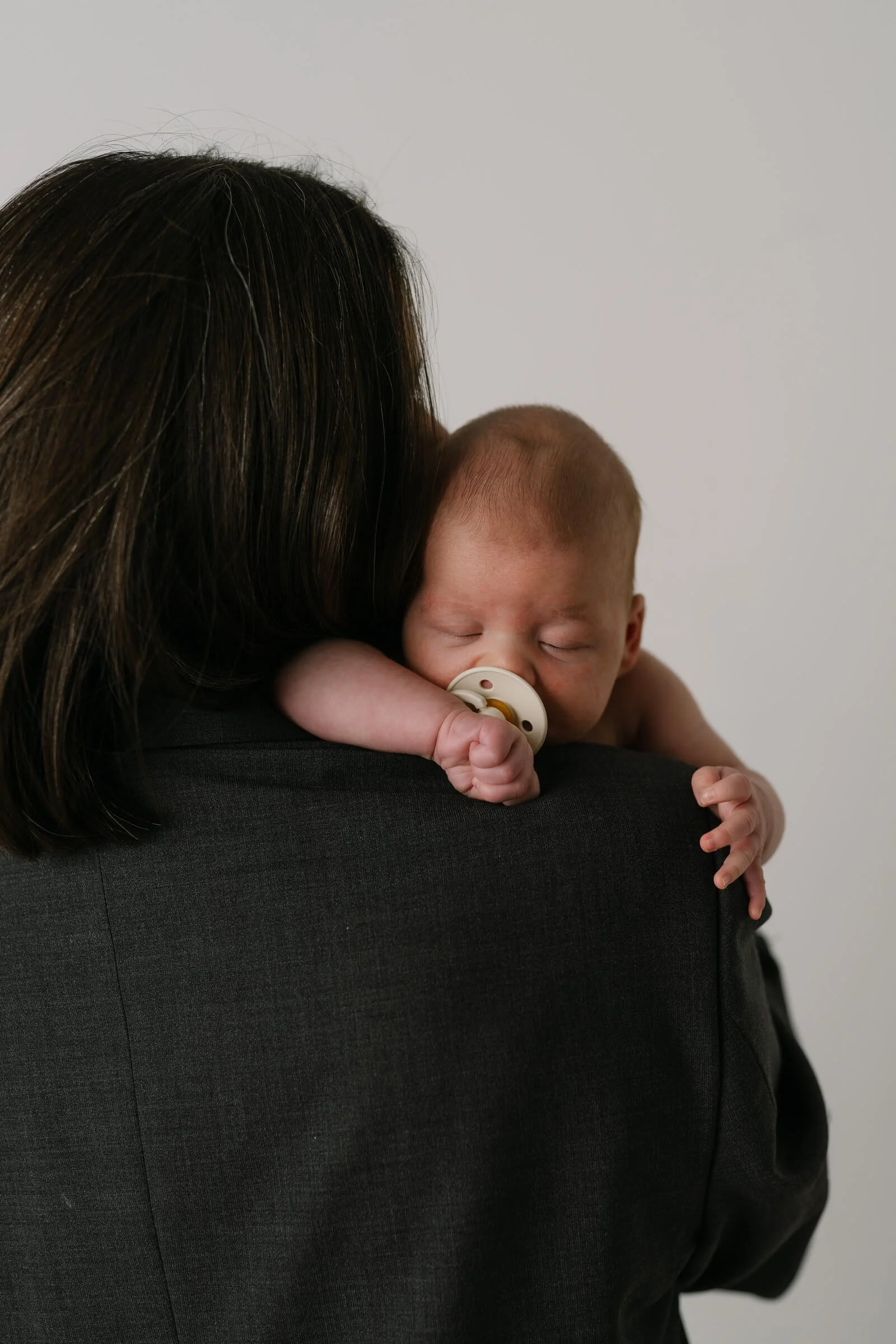 A woman holding a sleeping baby with a pacifier, seen from behind, against a plain white background.