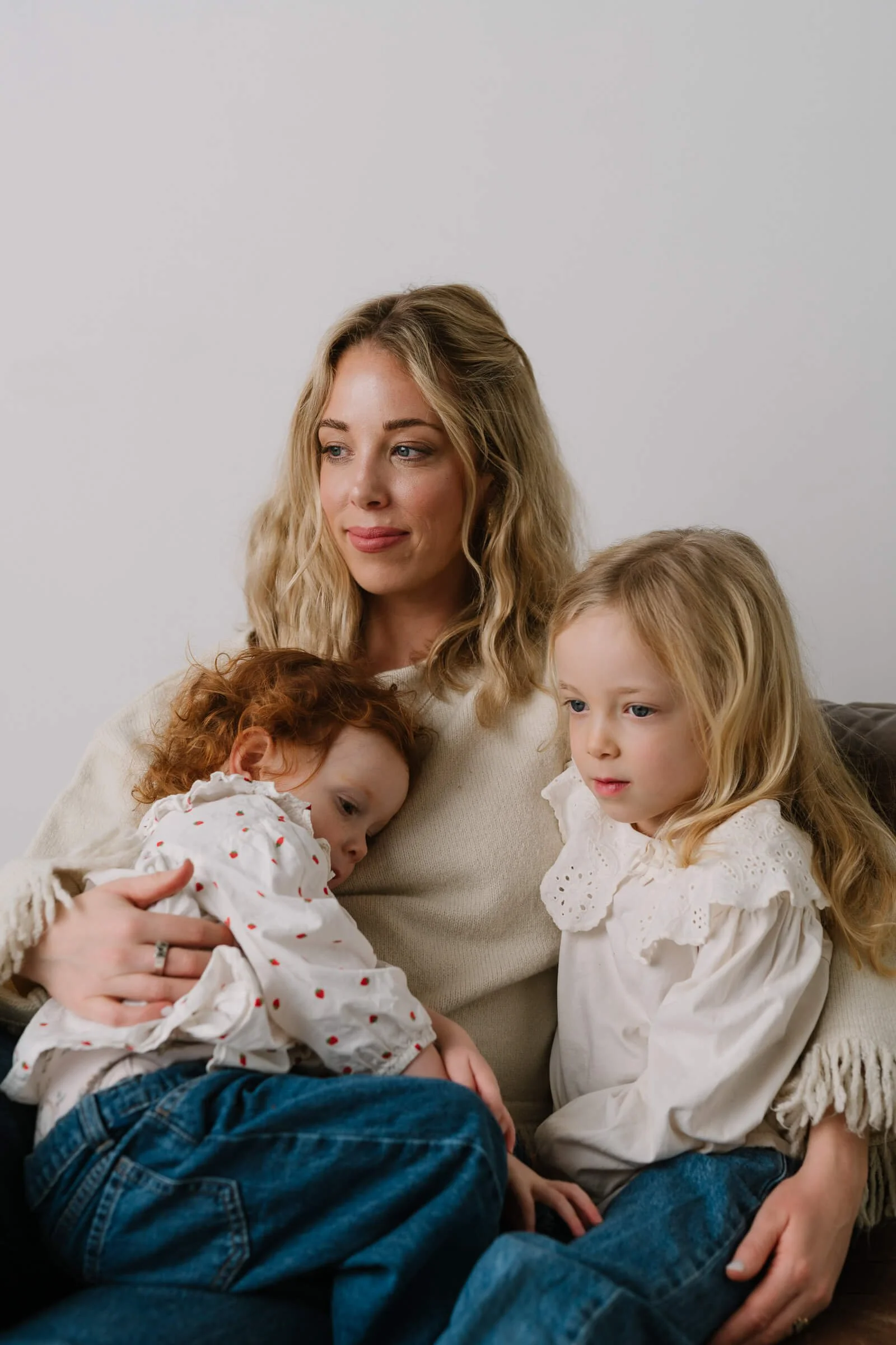 A woman with blonde hair sits on a couch holding two young girls, one with red curly hair resting her head on her lap and the other with blonde straight hair sitting beside her, all three children wearing white tops and jeans. The woman has a gentle 