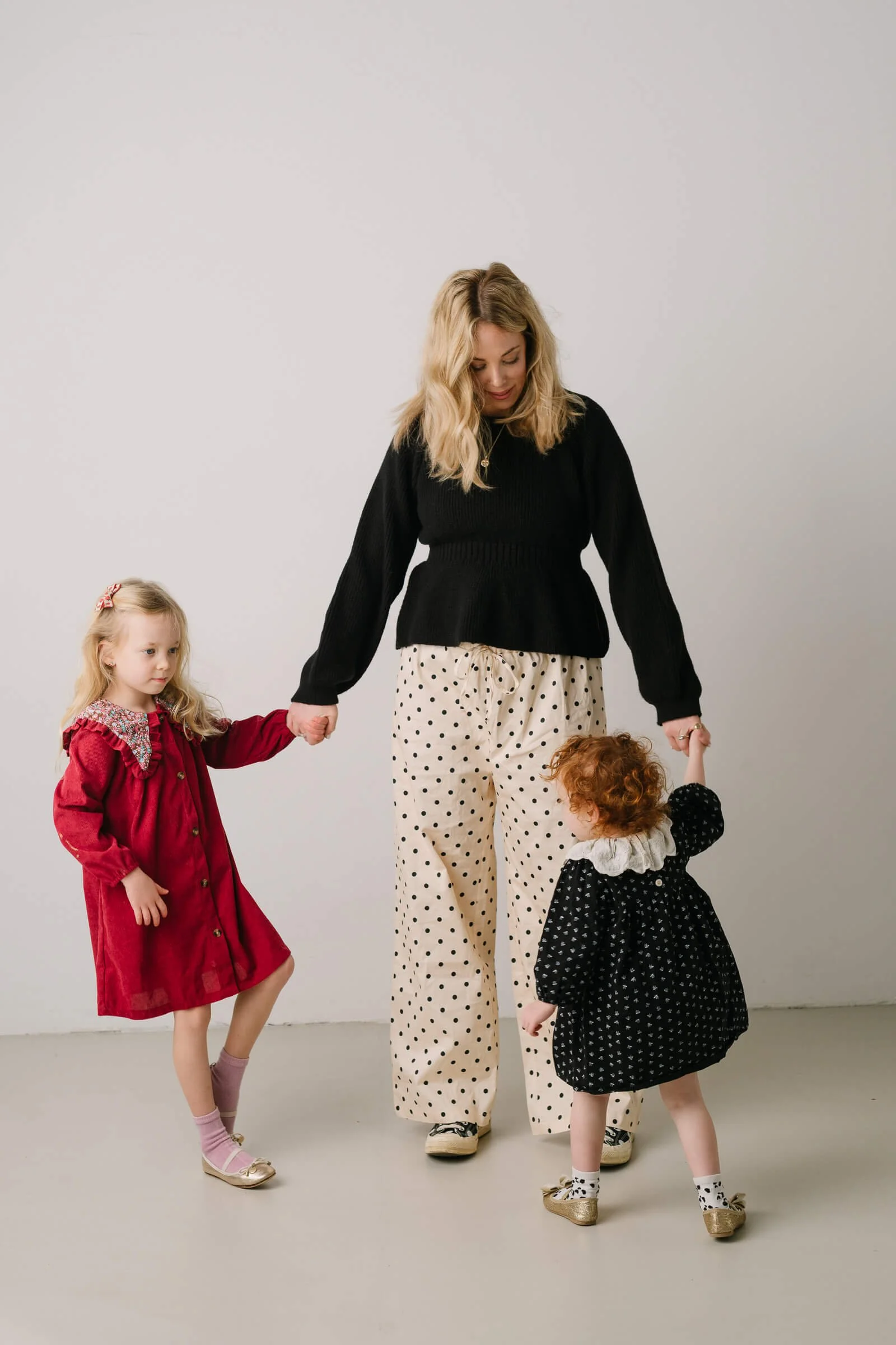 A woman walking with two young girls holding hands in a minimalistic studio with a white background.