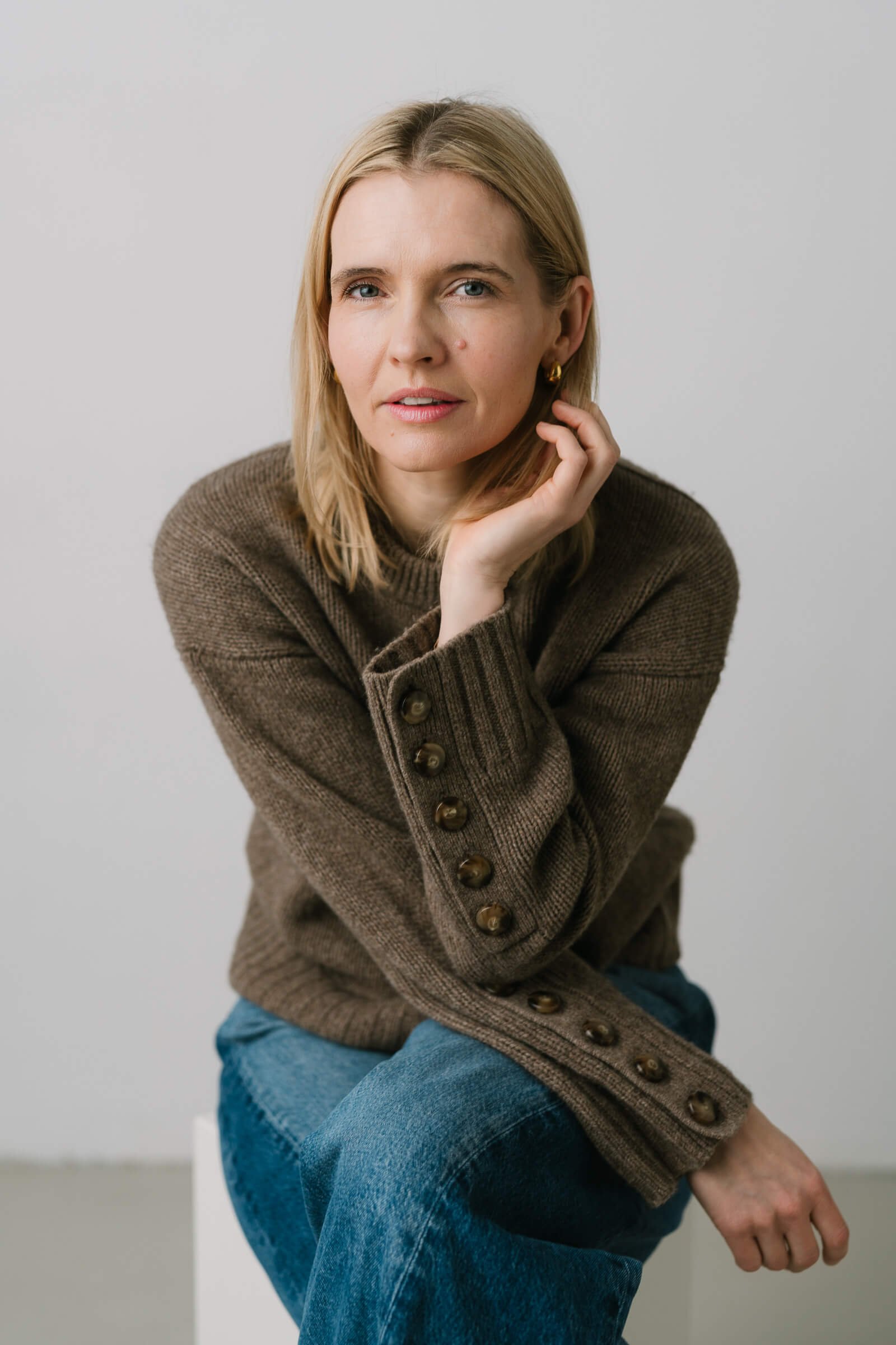 A woman with blonde hair, wearing a brown sweater with buttons on the sleeve, and blue jeans, sitting against a plain light grey background.