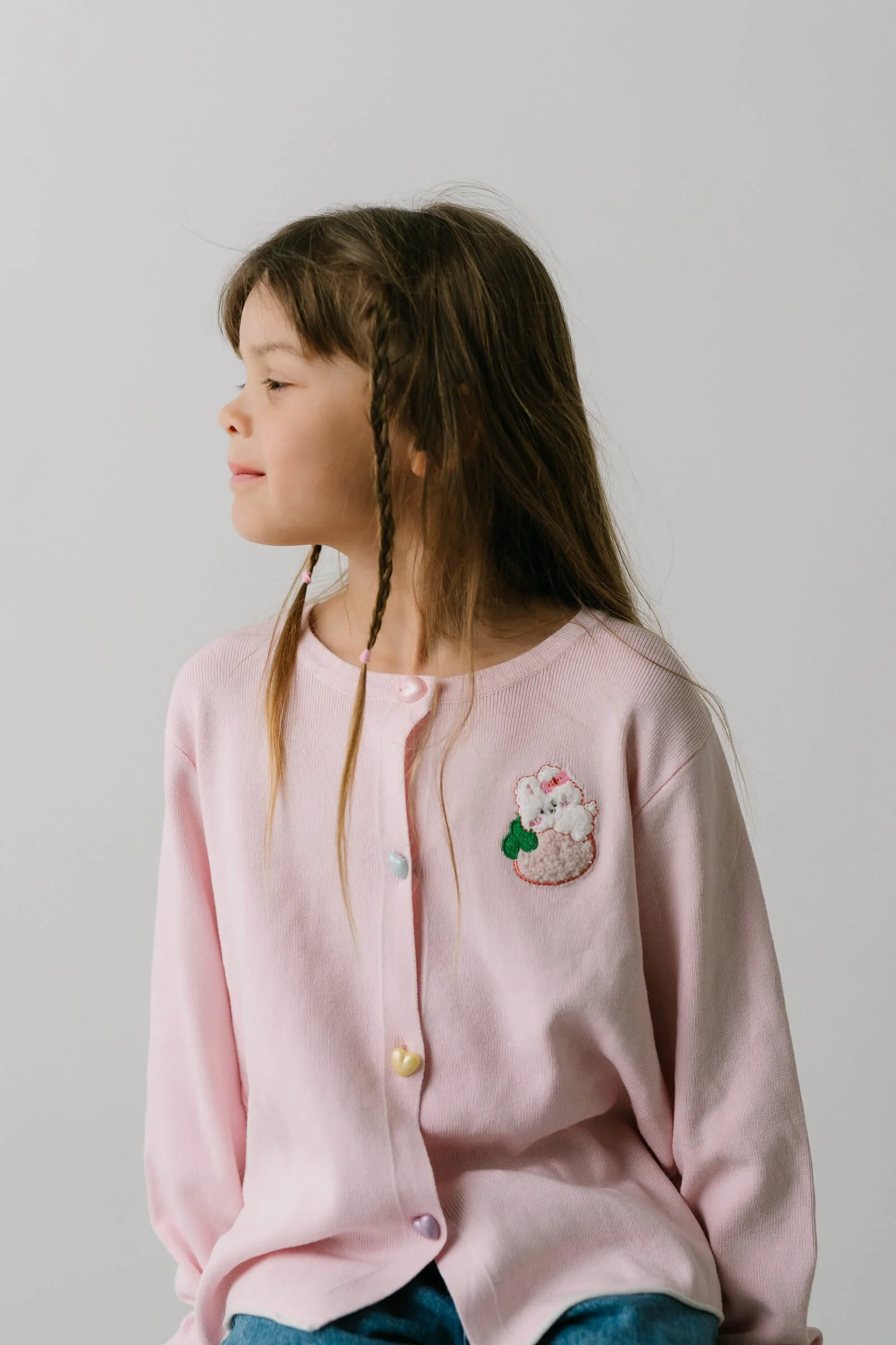 Young girl with long brown hair, some styled in small braids, wearing a pink cardigan with a cloud and cupcake embroidered patch, and colorful heart-shaped buttons.